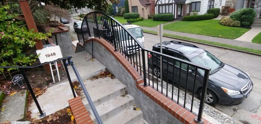 View of a black metal fence on a brick and concrete staircase leading to a sidewalk with parked cars and residential houses in the background.