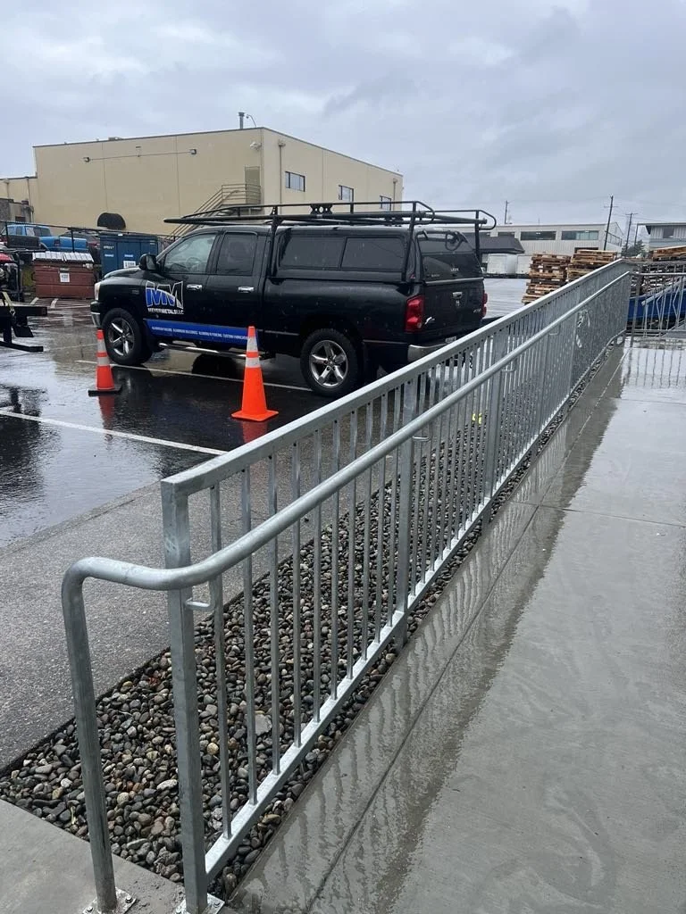 A black utility vehicle parked behind orange traffic cones on a rainy day, with a metal railing and pebbled landscaping in the foreground. Industrial warehouse buildings and a stack of pallets are visible in the background.