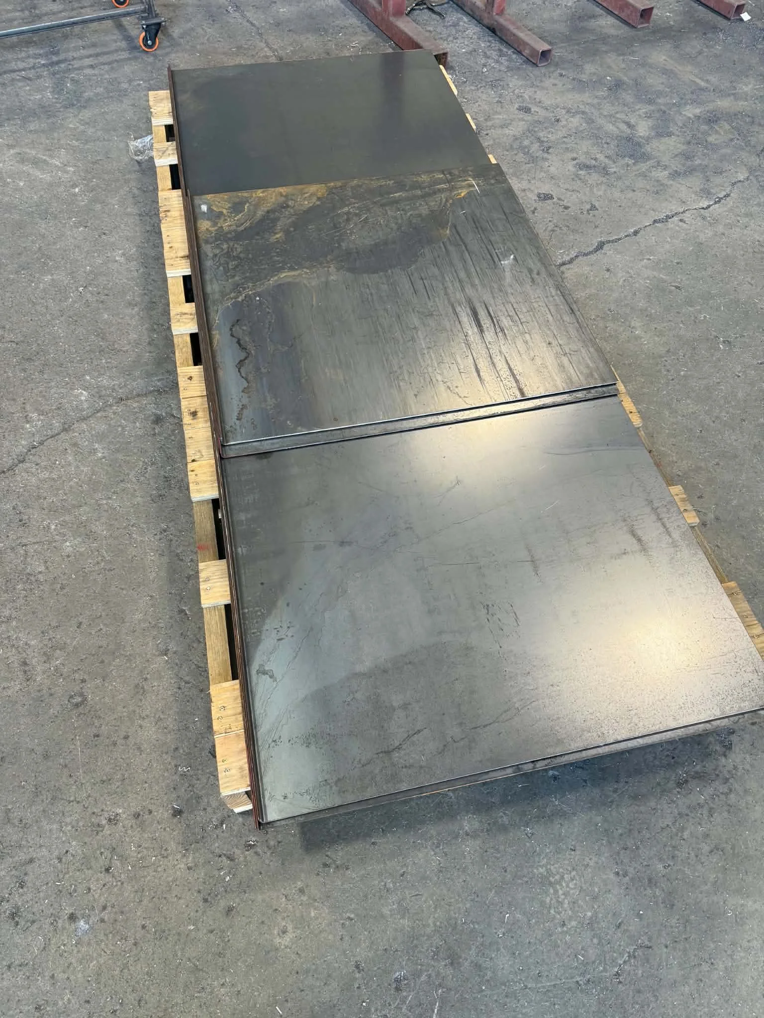 Four stainless steel sheets are on wooden pallets on a concrete floor, with metal pipes and tools in the background.