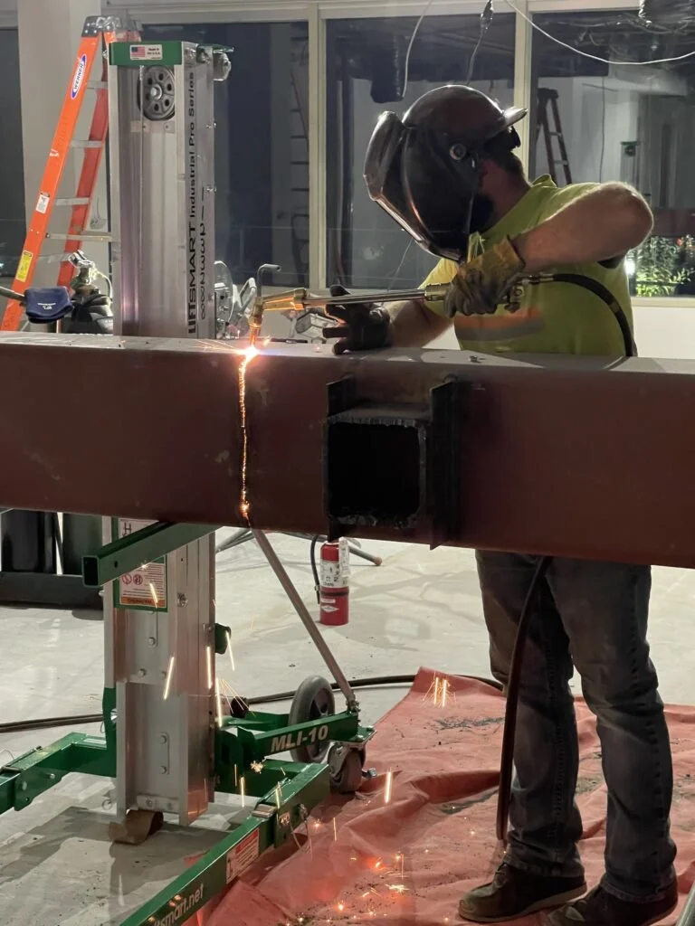 Worker welding a large metal beam using a protective helmet and gloves, with sparks flying during the welding process.