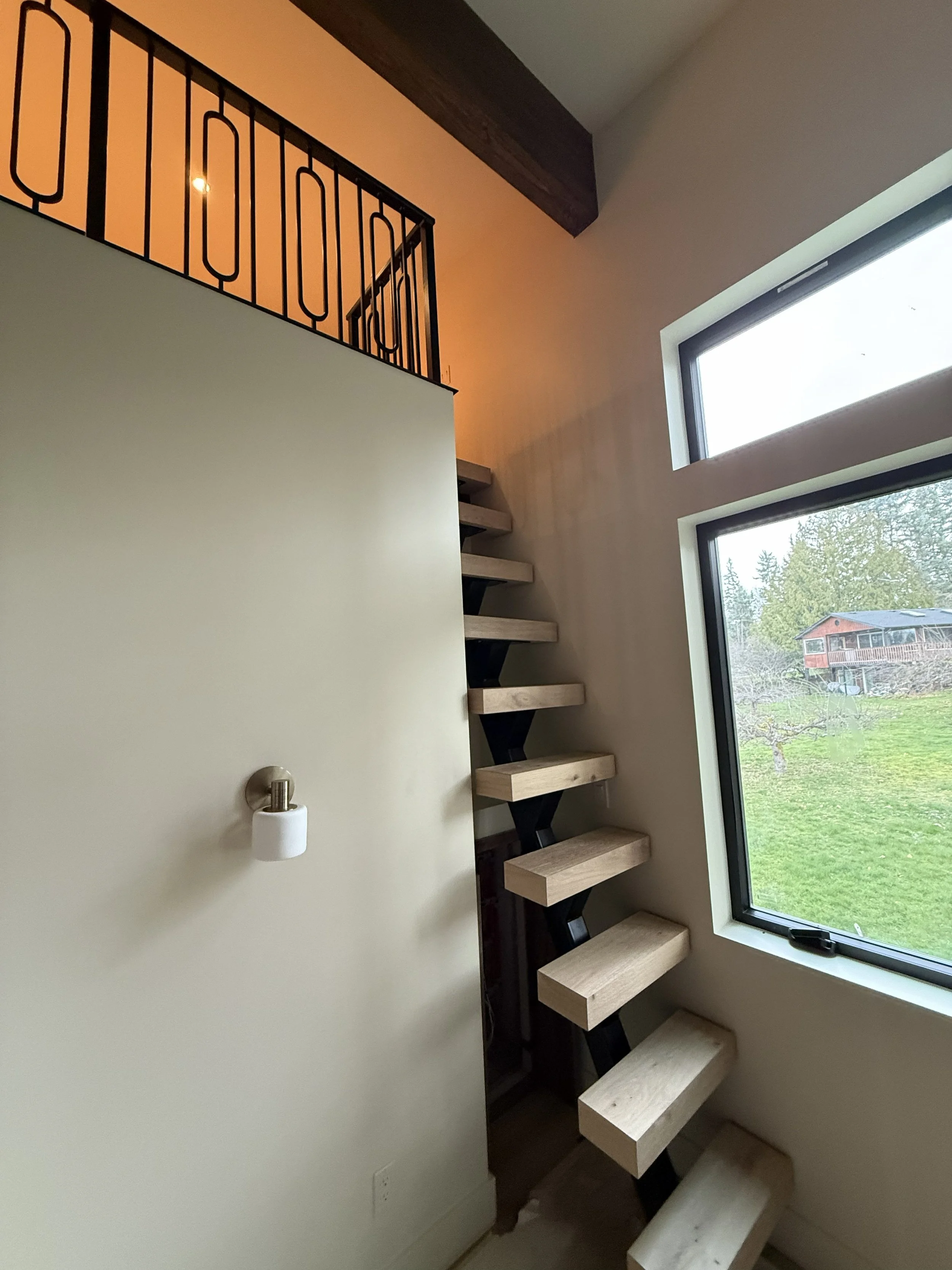 Interior view of a modern staircase with wooden treads and a black metal support structure, next to a large window showing a green outdoor landscape.