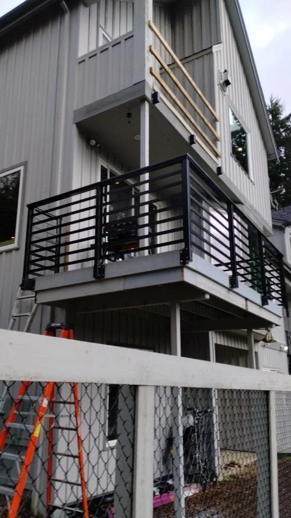 Outdoor view of a house with a second-floor balcony under construction, showing wooden railing supports, black metal balcony railing, and tools like ladders and bikes below.