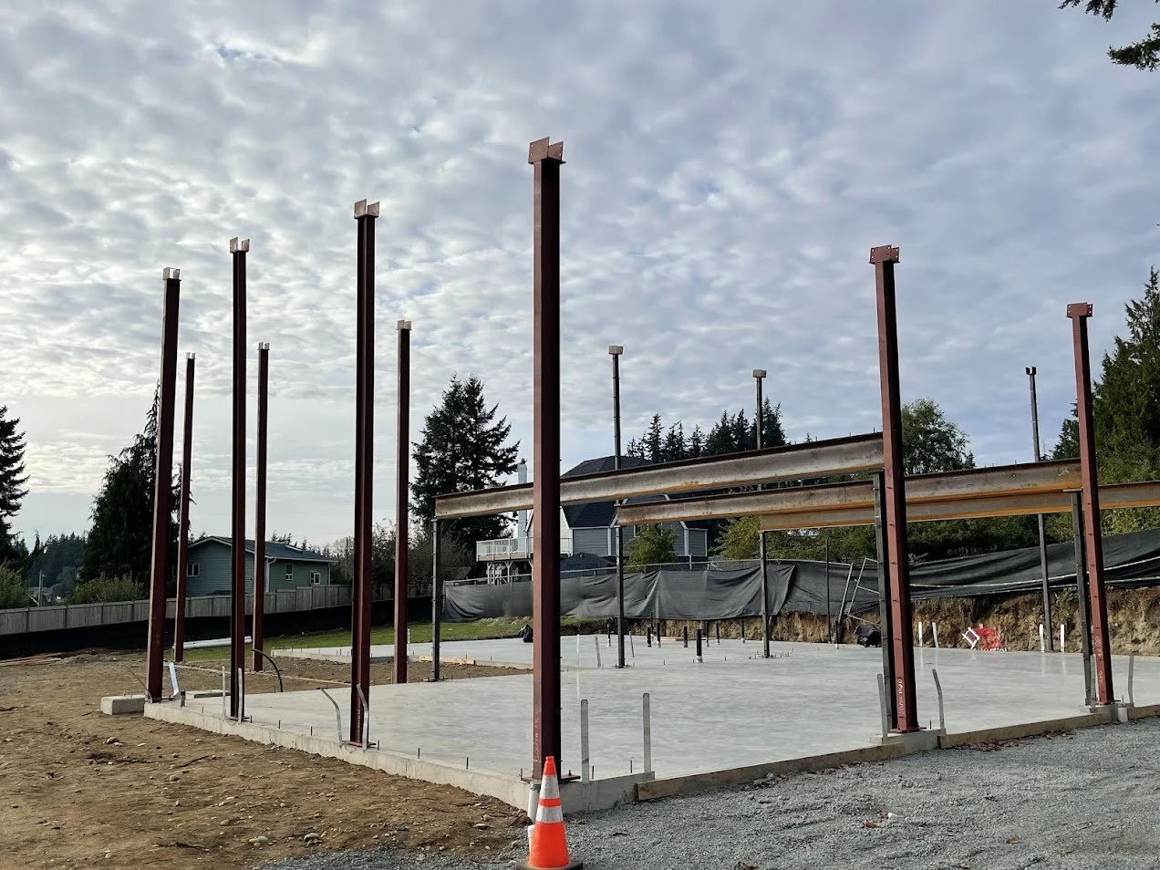 Under construction building foundation with steel beams and concrete slab, orange traffic cone, surrounded by residential houses and trees under partly cloudy sky.