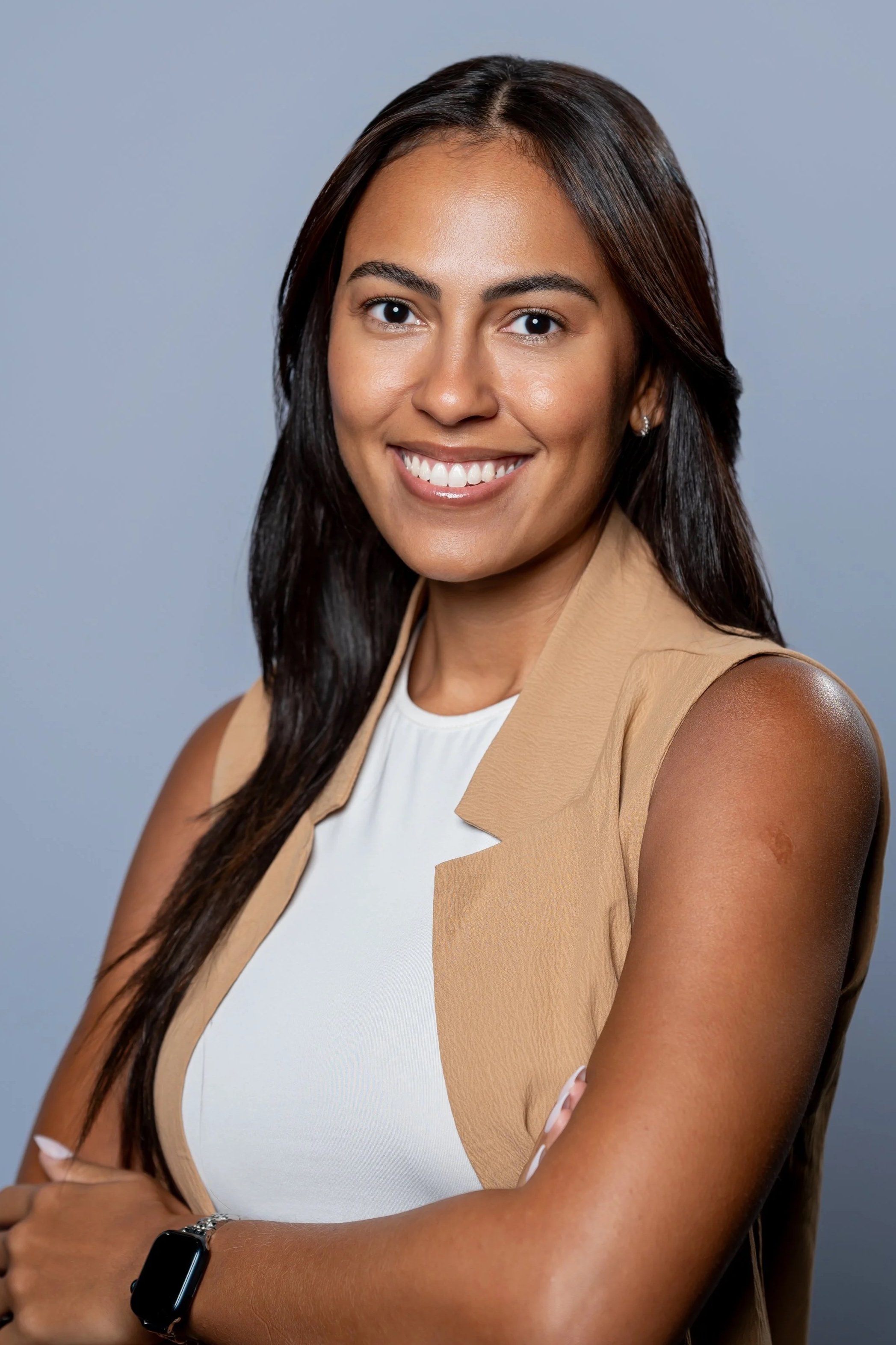 A woman with long dark hair, wearing a sleeveless beige and white top, smiling with her arms crossed, against a gray background.