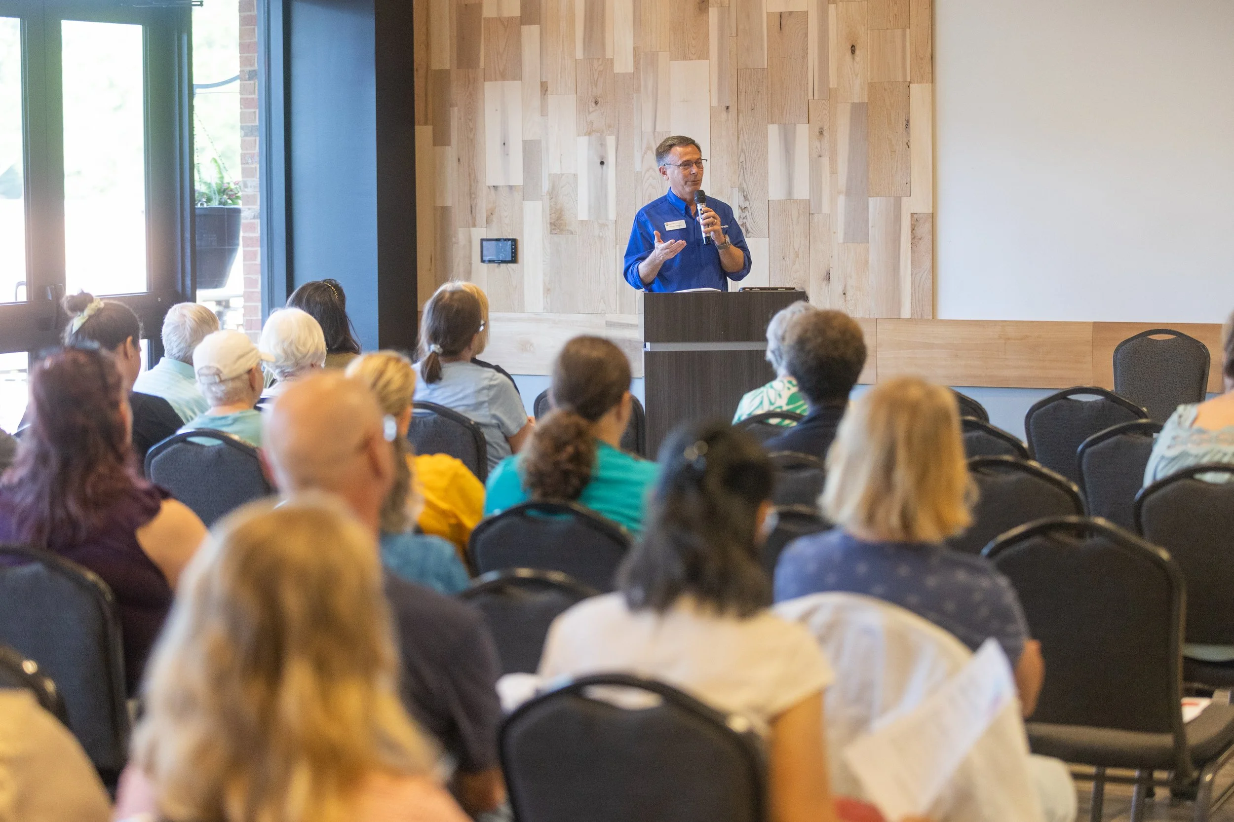 A man in a blue shirt giving a presentation to an audience in a room with a wood-paneled wall and a large window.