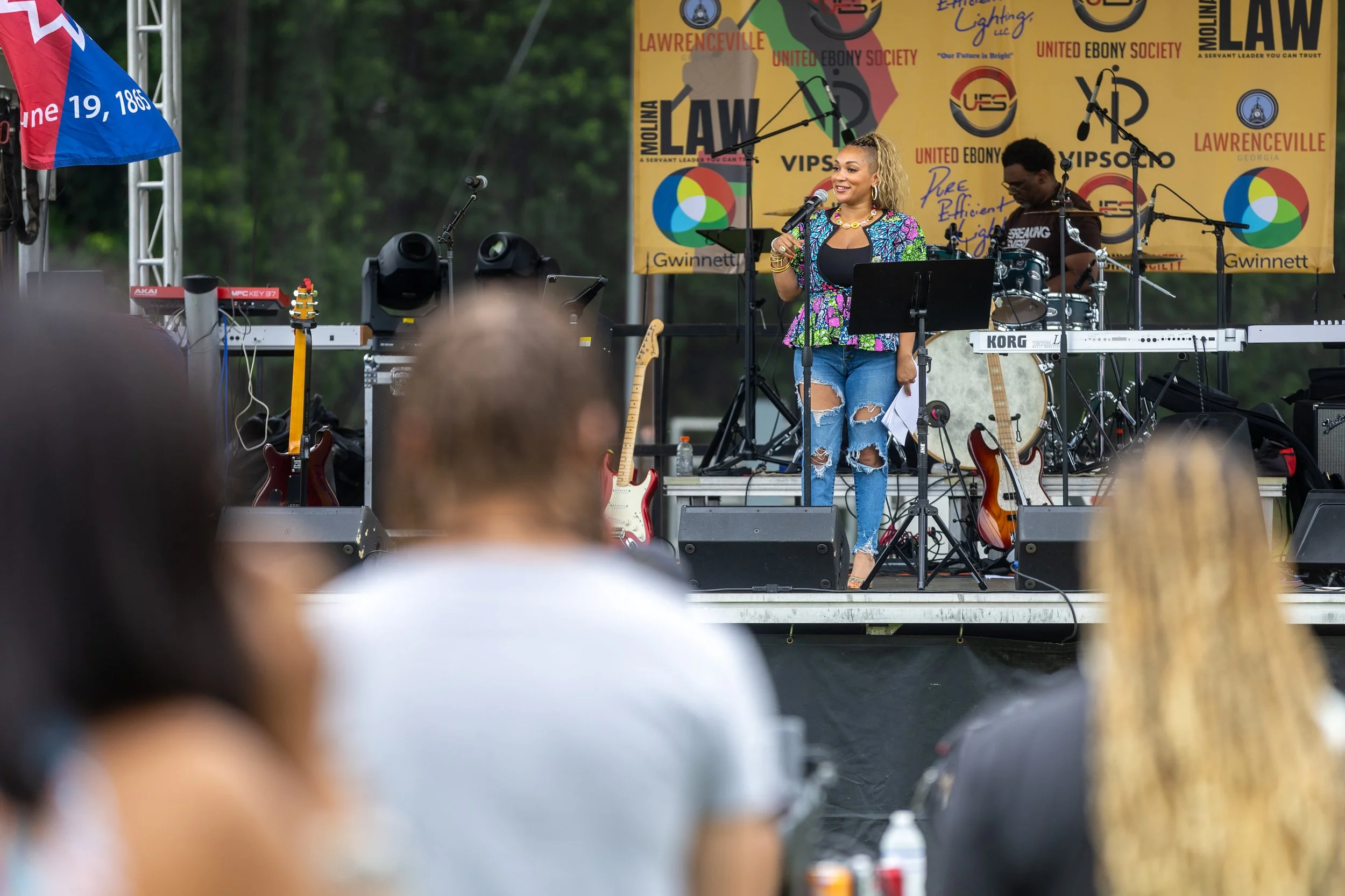 A woman with blonde hair singing into a microphone on stage at an outdoor event, with a drummer and musical equipment behind her, and a large yellow banner with logos and text in the background.