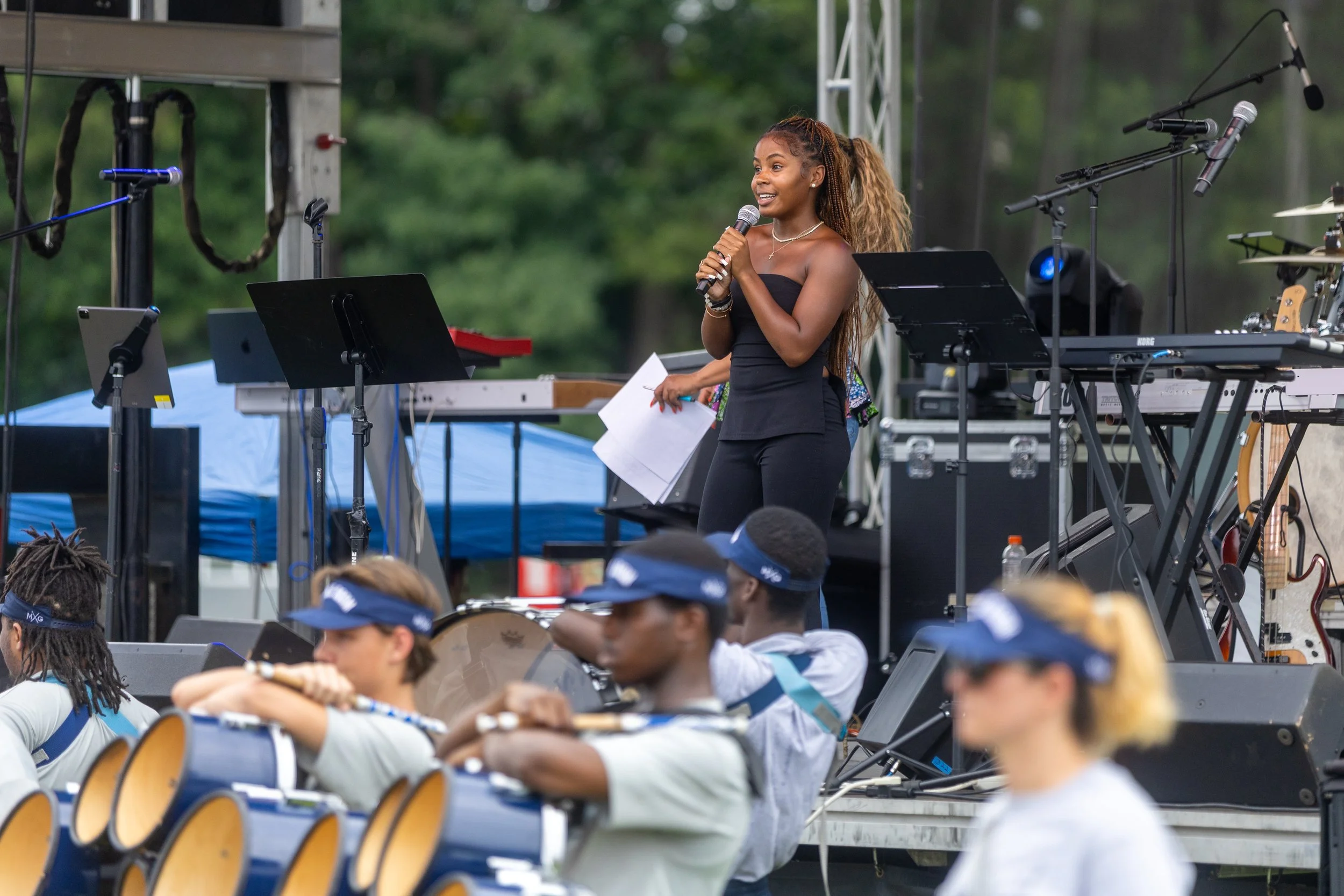 A woman speaking into a microphone on an outdoor stage surrounded by musical equipment and audience members wearing hats.