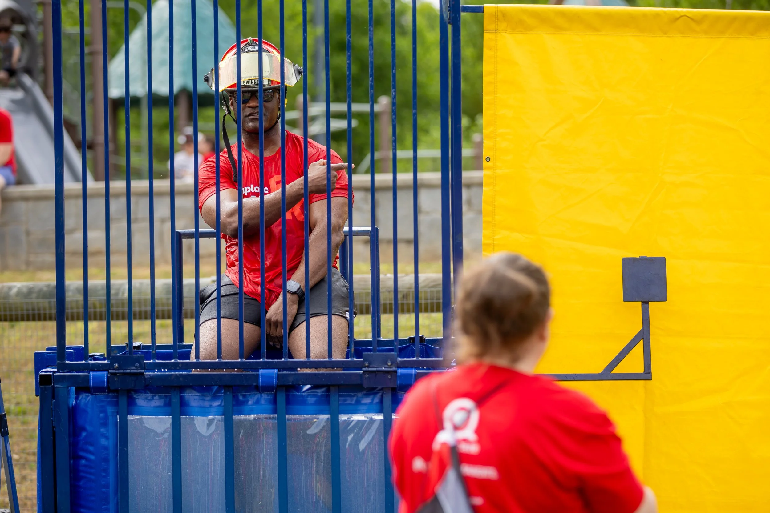 A man sitting in a cage, wearing a firefighter helmet and sunglasses, pointing to the right. A boy in a red shirt stands in front, with the back of his head visible, at an outdoor event.