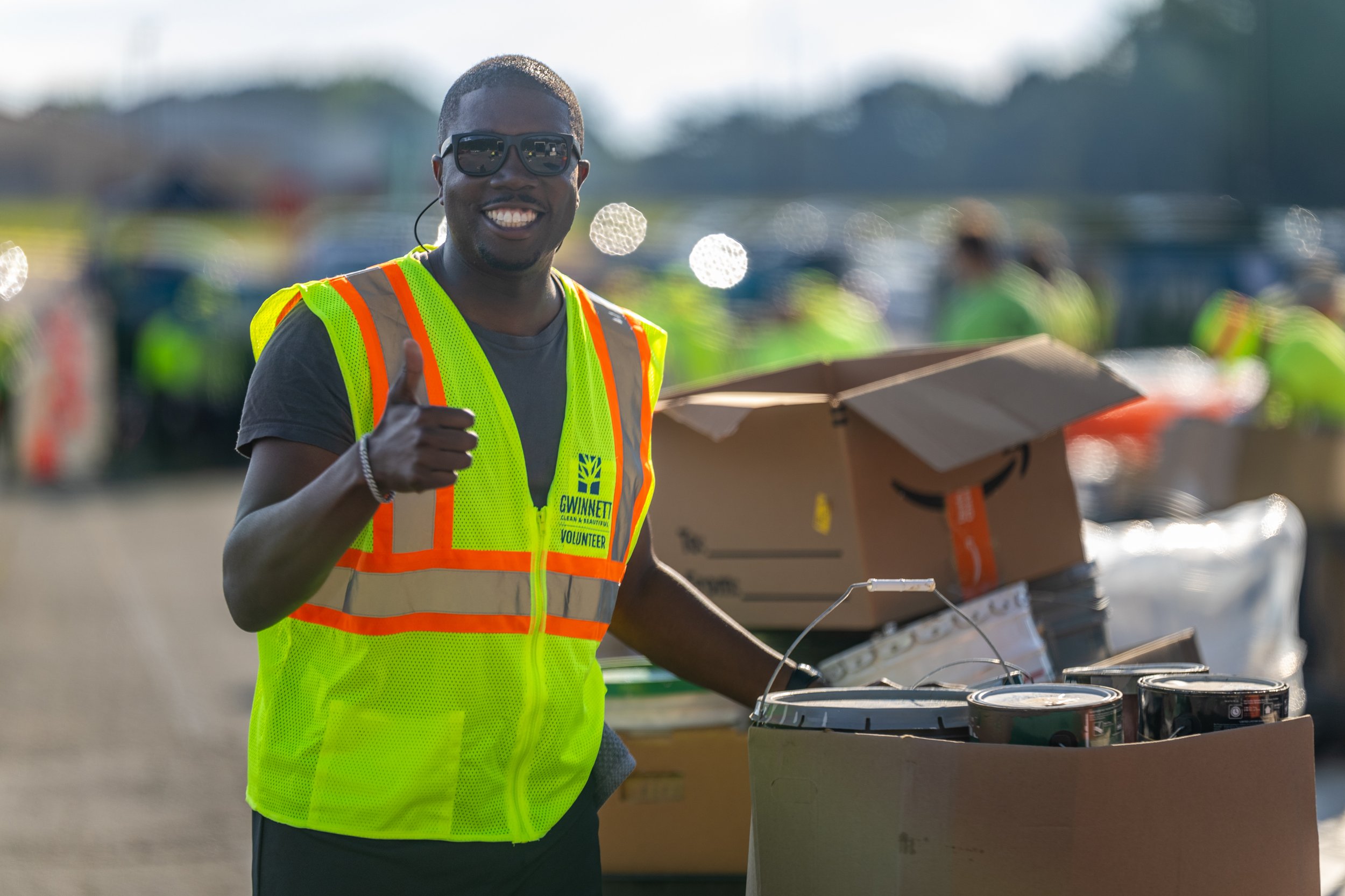 Smiling man wearing sunglasses and a bright safety vest with 'GWINNETT VOLUNTEER' on it, standing outdoors among other volunteers, and holding a box of canned goods.