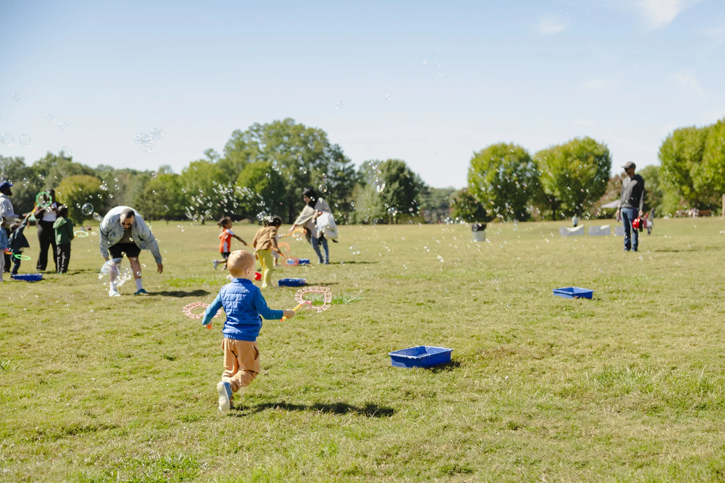 Children and adults playing with bubbles in a park on a sunny day, with green trees and blue sky in the background.