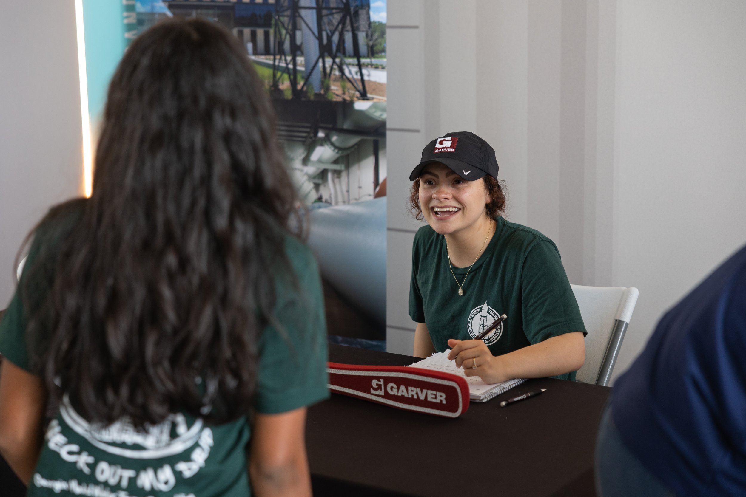 A woman with curly hair, wearing a black cap and a green shirt, is sitting at a table with a group of people in an indoor setting, smiling and talking. There are notebooks and pens on the table, and a large screen or television in the background.