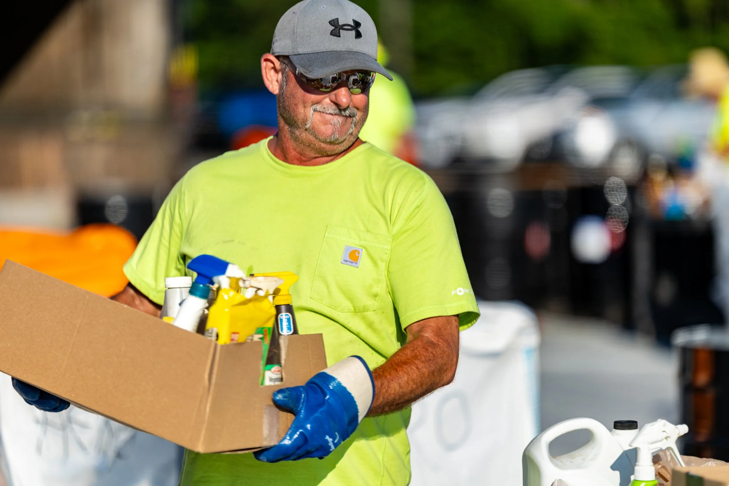A man in a gray Under Armour cap, black sunglasses, a bright yellow Carhartt t-shirt, and blue gloves is participating in a community cleanup event. He is holding a cardboard box filled with spray bottles and cleaning supplies, standing outdoors with blurred cars and greenery in the background.