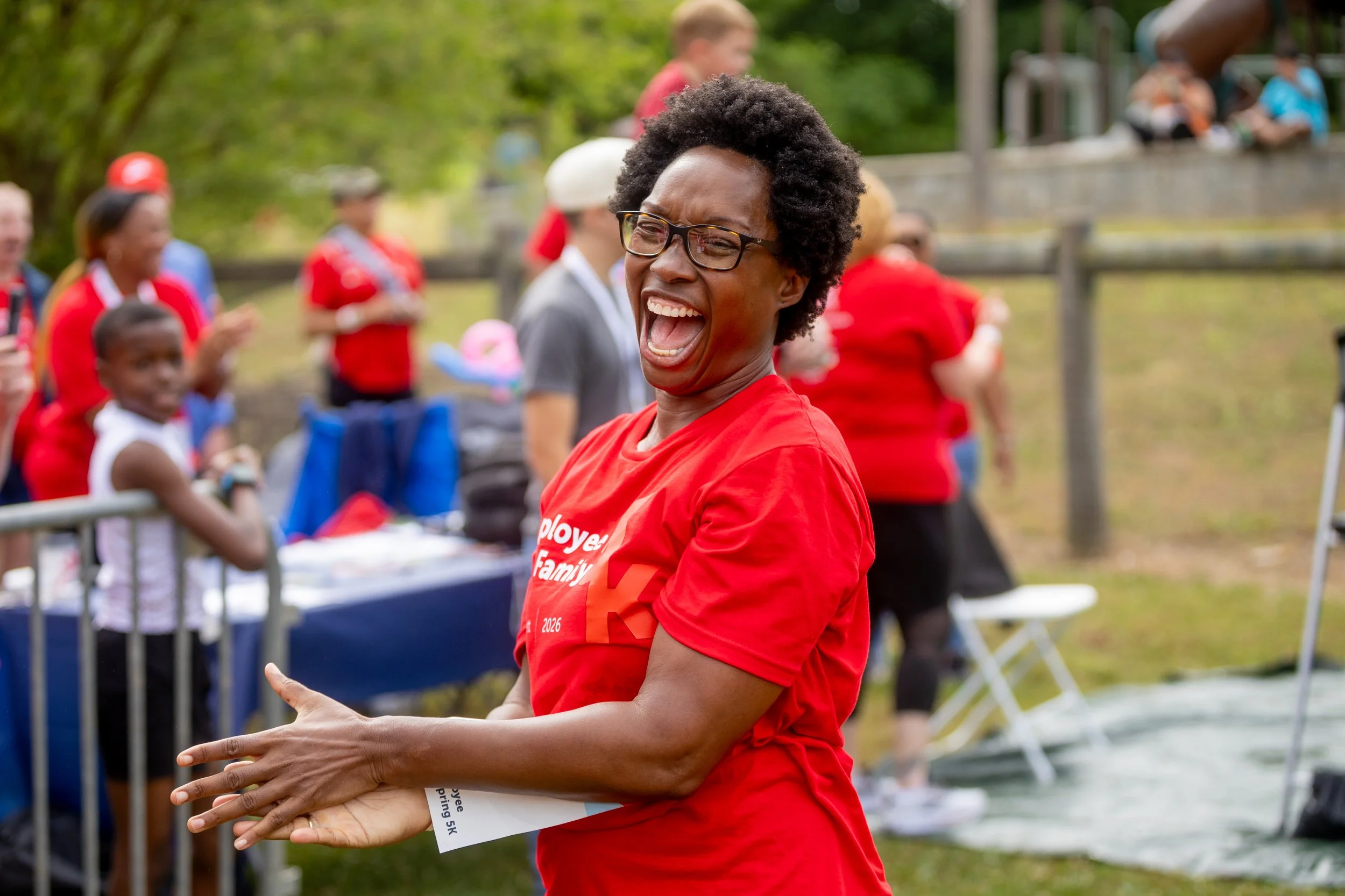Woman with glasses and curly hair, wearing a red shirt, smiling and clapping at an outdoor event with trees and people in the background.