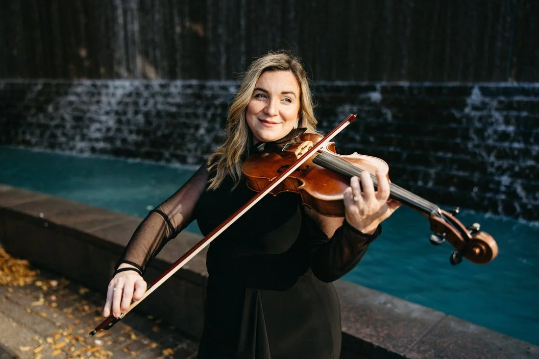 A woman with blonde hair playing the violin beside a pool with a waterfall in the background.