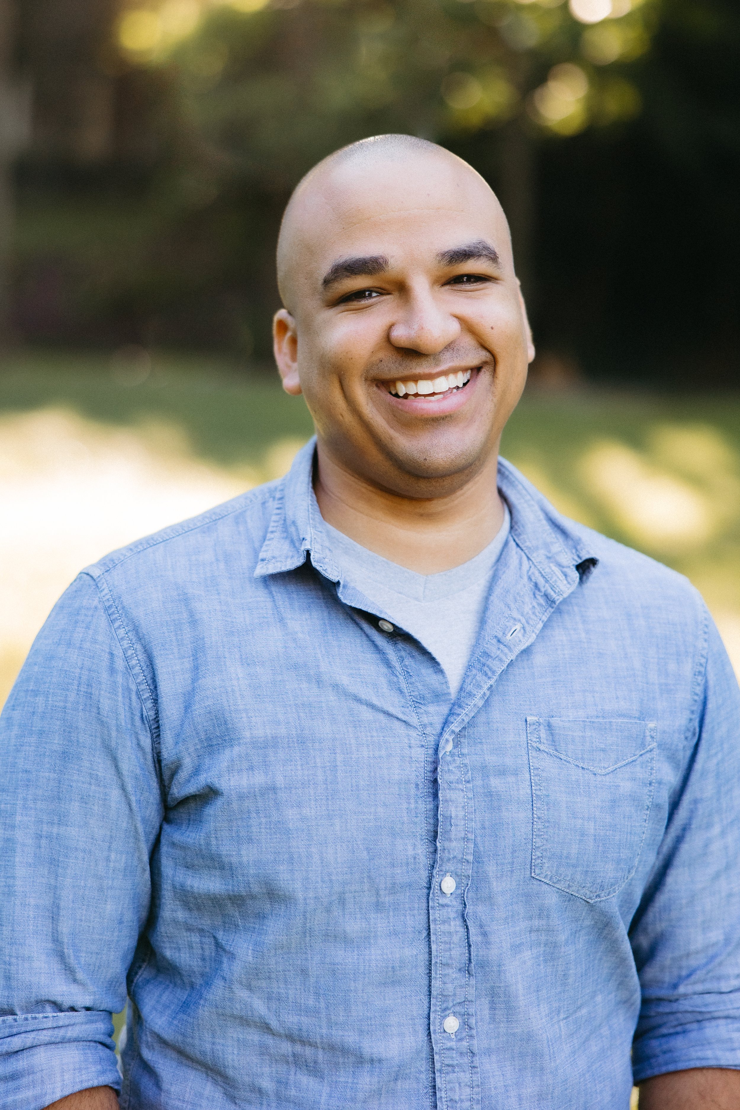 A smiling man with a bald head, wearing a light blue button-up shirt, standing outdoors with blurred green trees in the background.