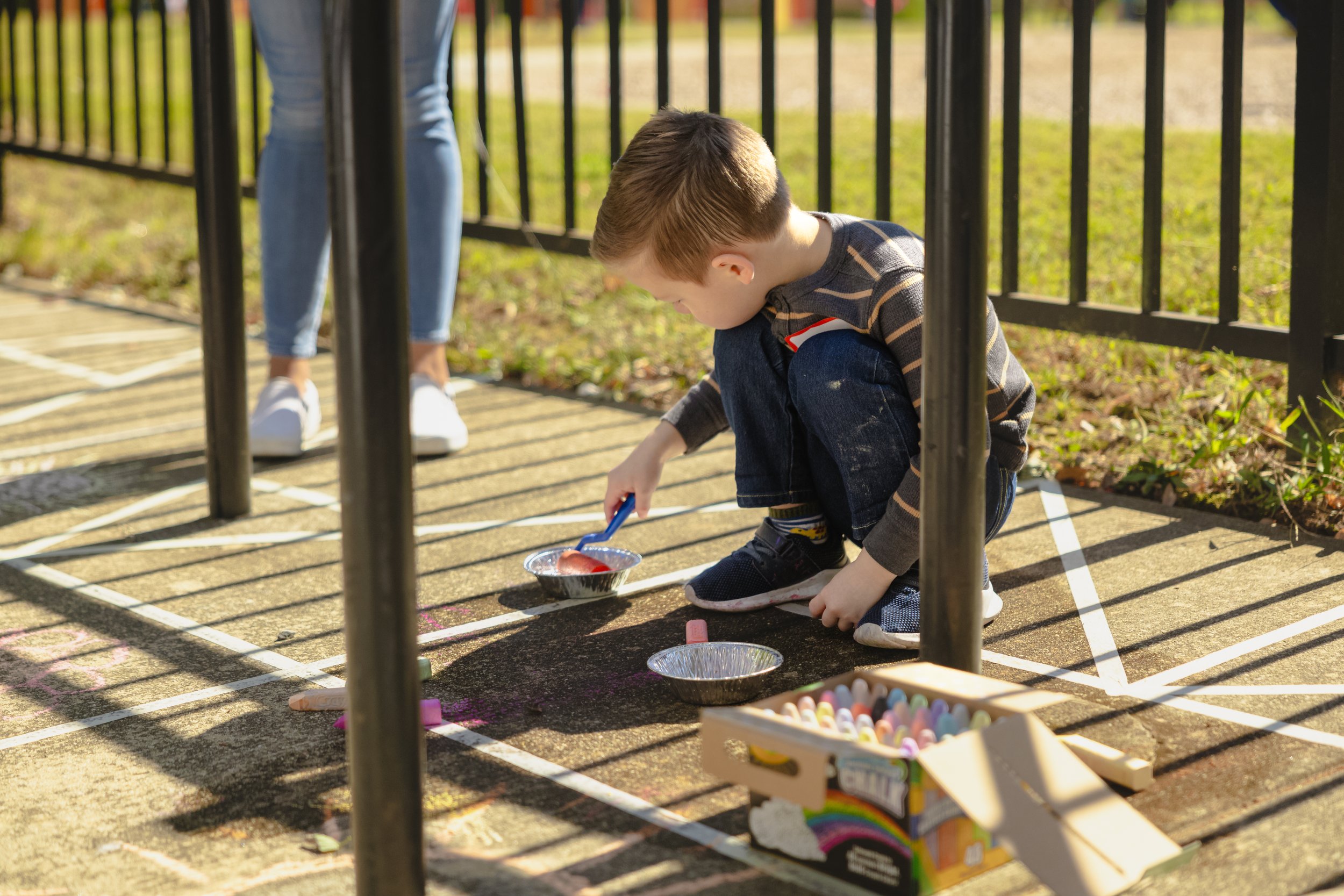 A young boy kneeling on the ground, drawing with chalk inside a fenced outdoor area, with colorful chalk and art supplies nearby.