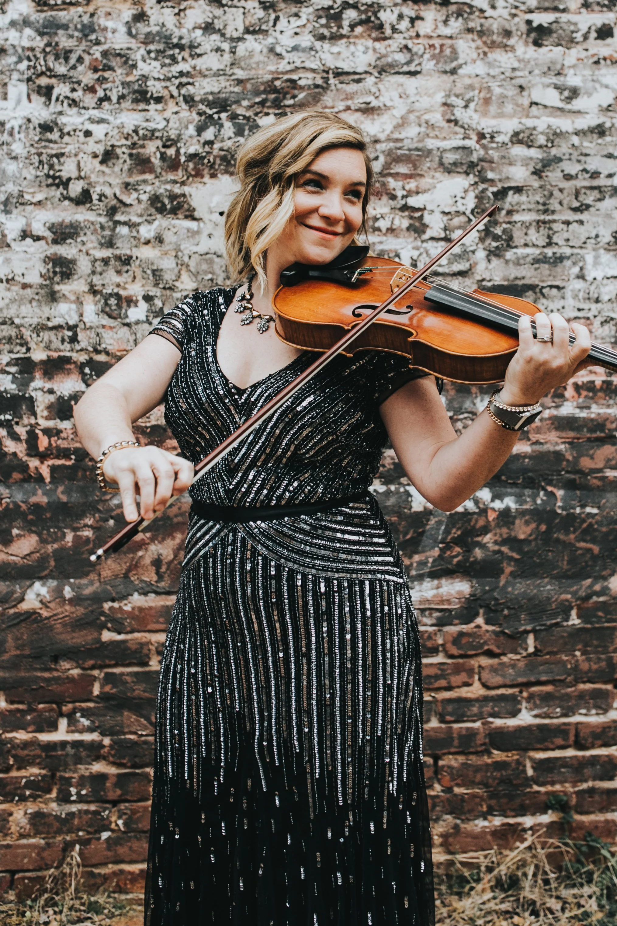 A woman with shoulder-length blonde hair playing a violin against a brick wall background.