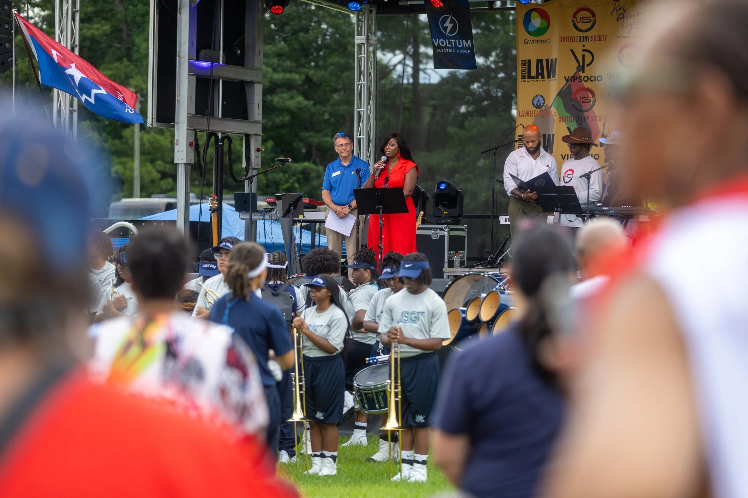 A stage at an outdoor event with people speaking and performing music, surrounded by a crowd of attendees, some in band uniforms holding musical instruments, and banners and flags in the background.