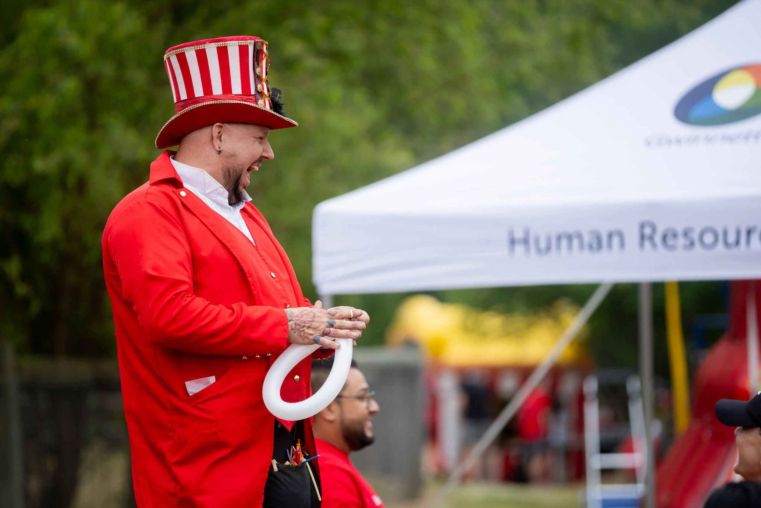 A man wearing a red jacket and a tall, red and white striped hat with gold accents, smiling with eyes closed, standing outdoors with a blurred crowd and a white tent with blue and yellow logo in the background.