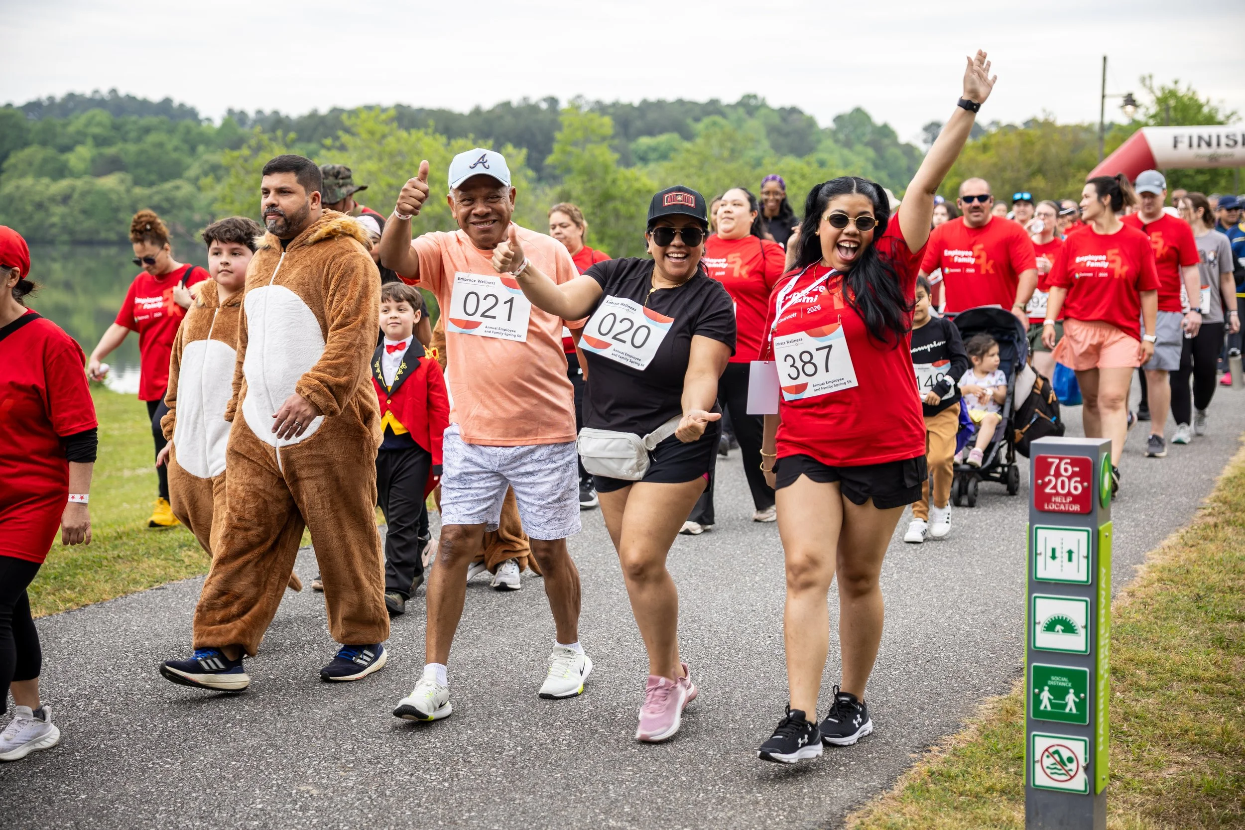 People participating in a community walk or run event outdoors, some dressed in costumes, including a person in a bear costume, smiling and waving near a finish line in a park with green trees in the background.