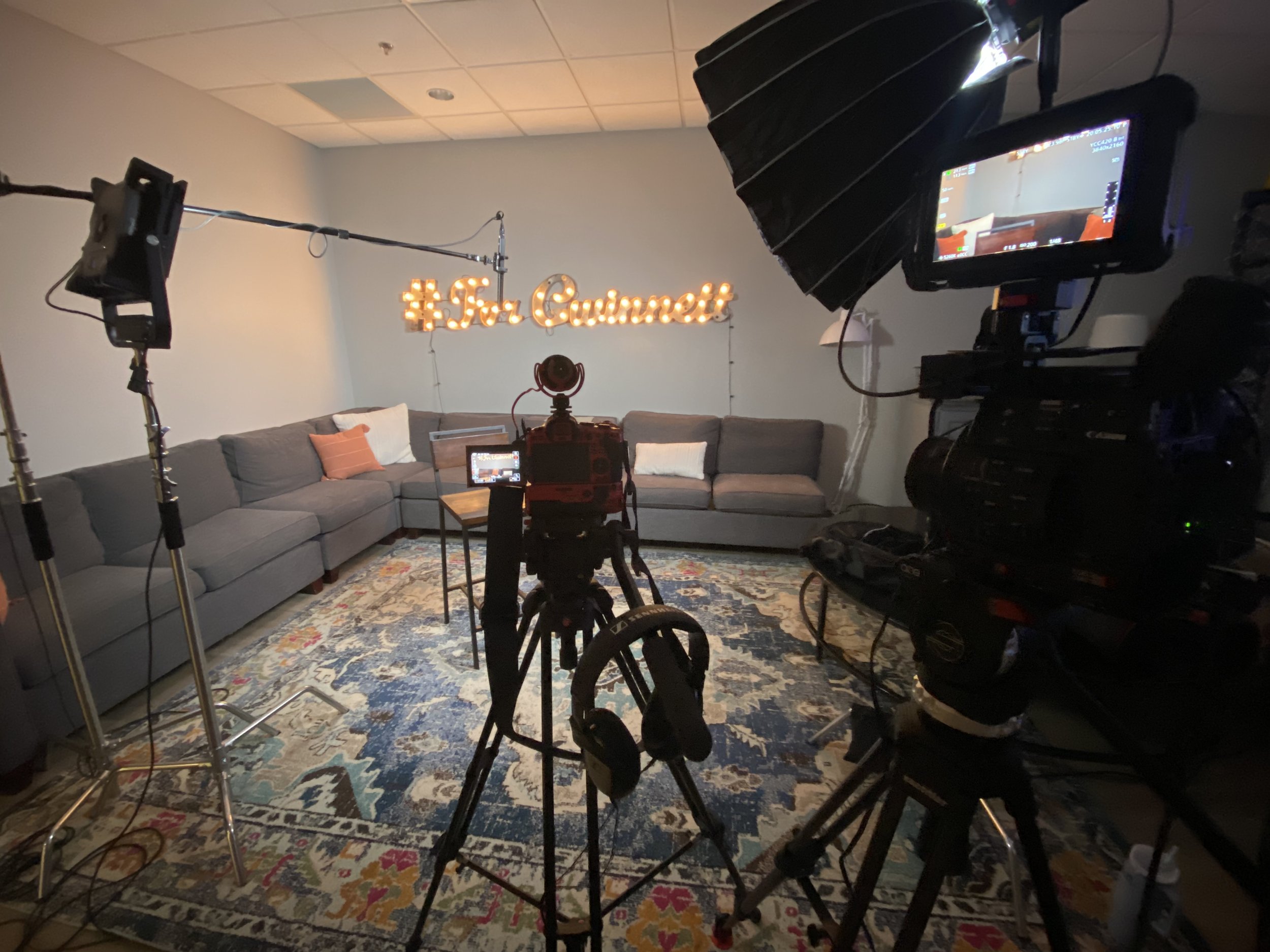A television or monitor on a light-colored wall displaying a view of a studio or interview setup with various film equipment and lighting. There is a gray sectional sofa with pillows, a patterned rug, and a hashtag sign that reads #ForCummett on the wall.