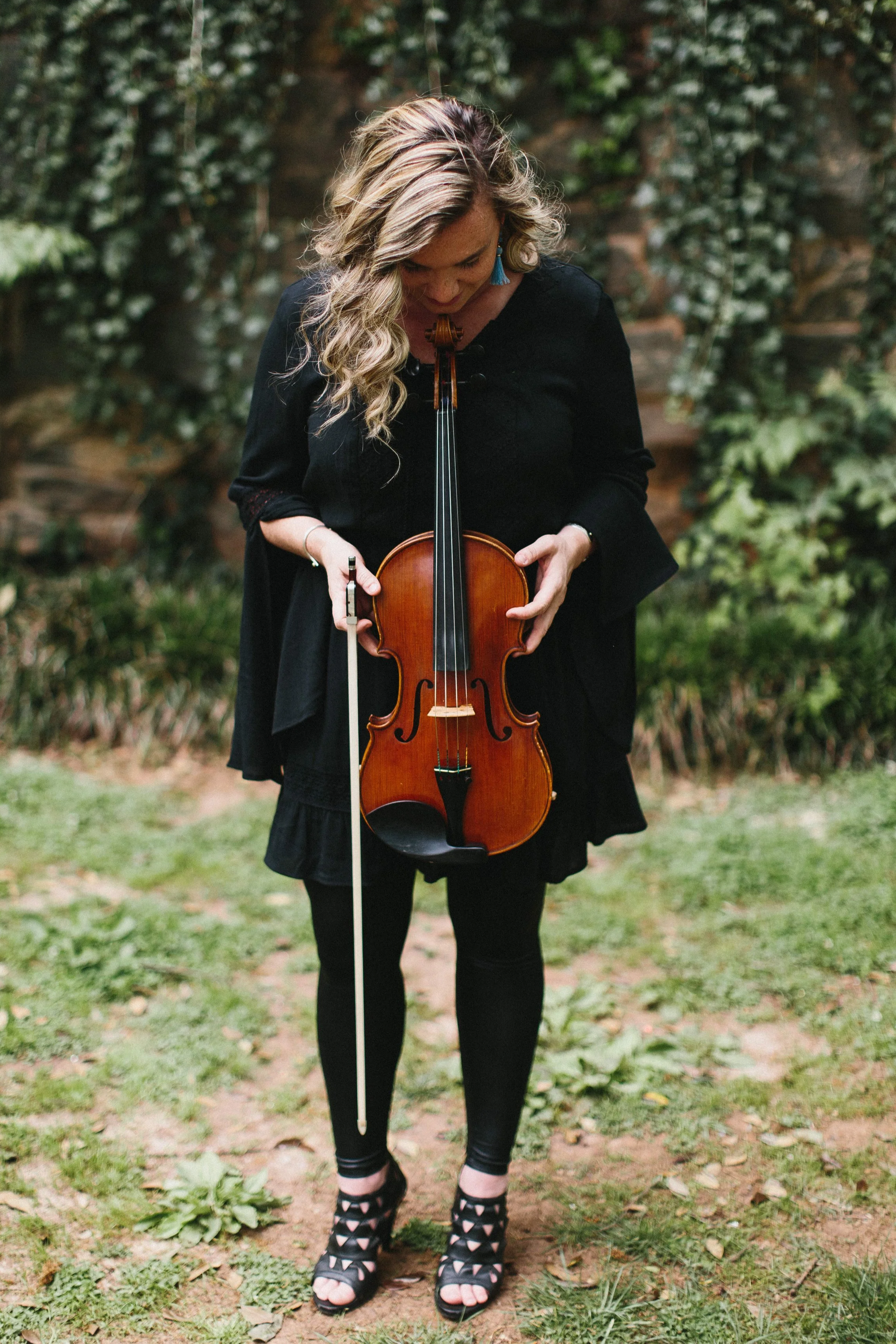 Woman with blonde, wavy hair looking down while holding a violin, outdoors in a garden with green foliage.