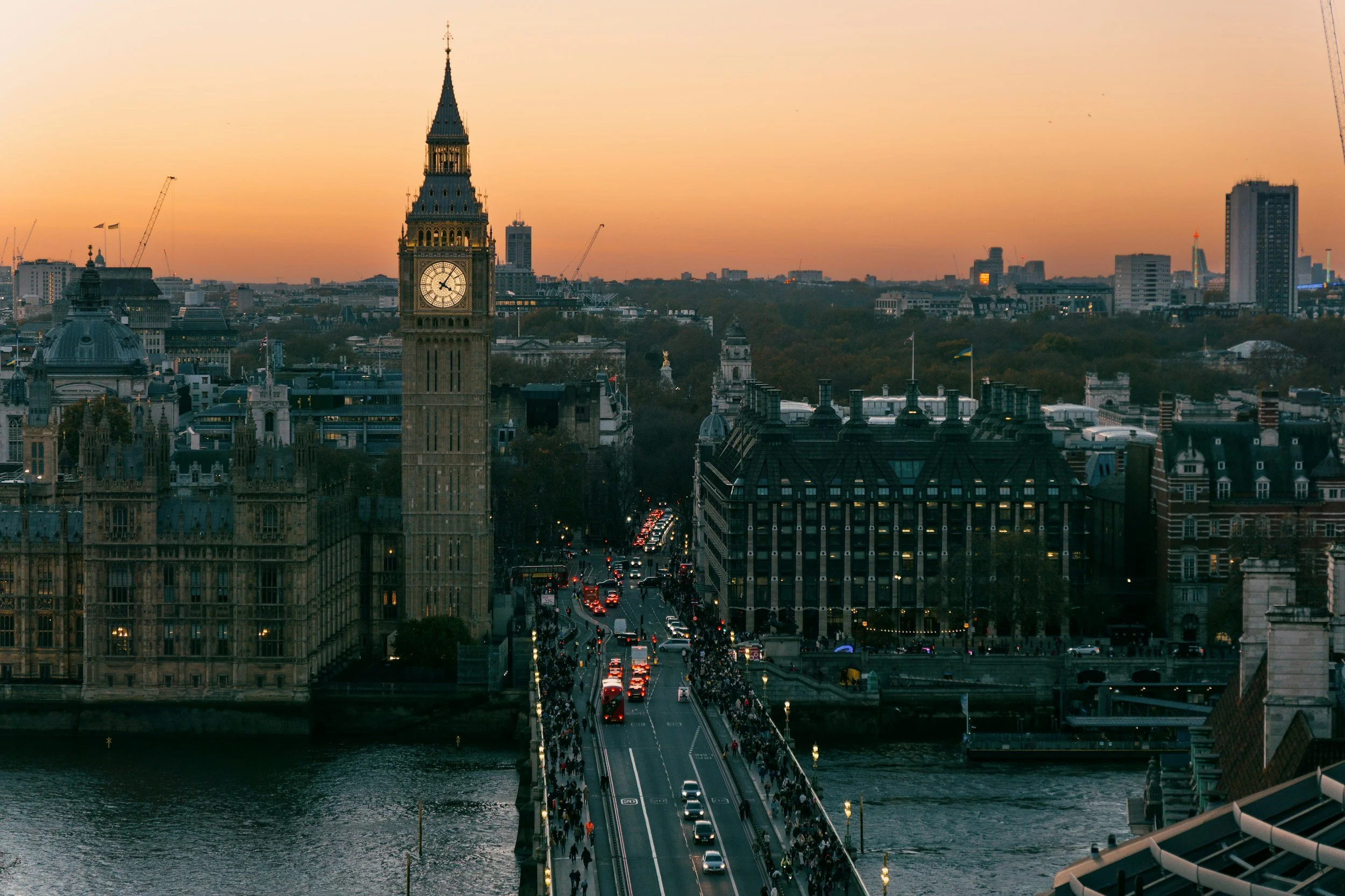 A cityscape of London featuring the Big Ben clock tower, with a bridge over a river and buildings in the background during sunset.