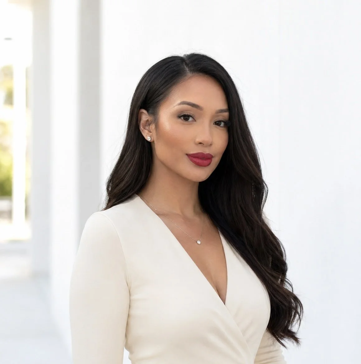 Portrait of a woman with long dark hair, wearing a white top, earrings, and a necklace, standing in a bright room with white background.