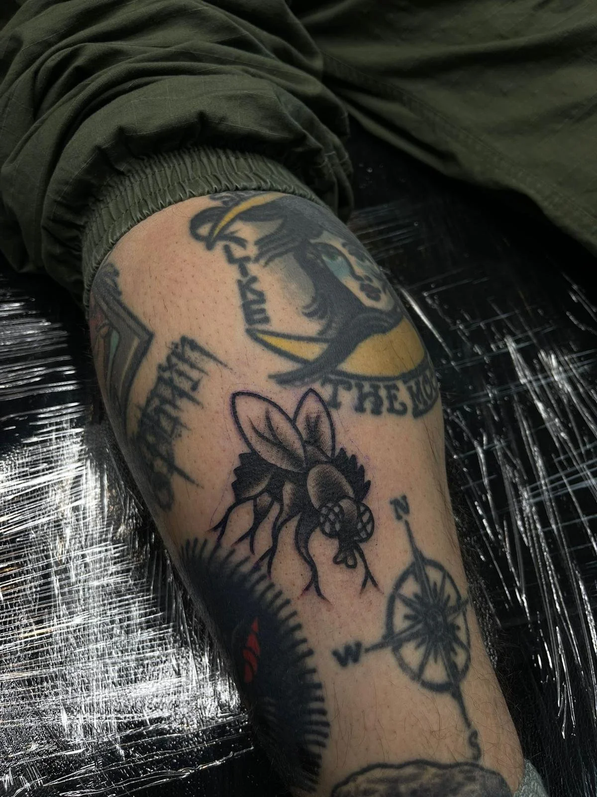 A person's upper arm with several tattoos including a bee, a compass, and a circular emblem with a bird and text.