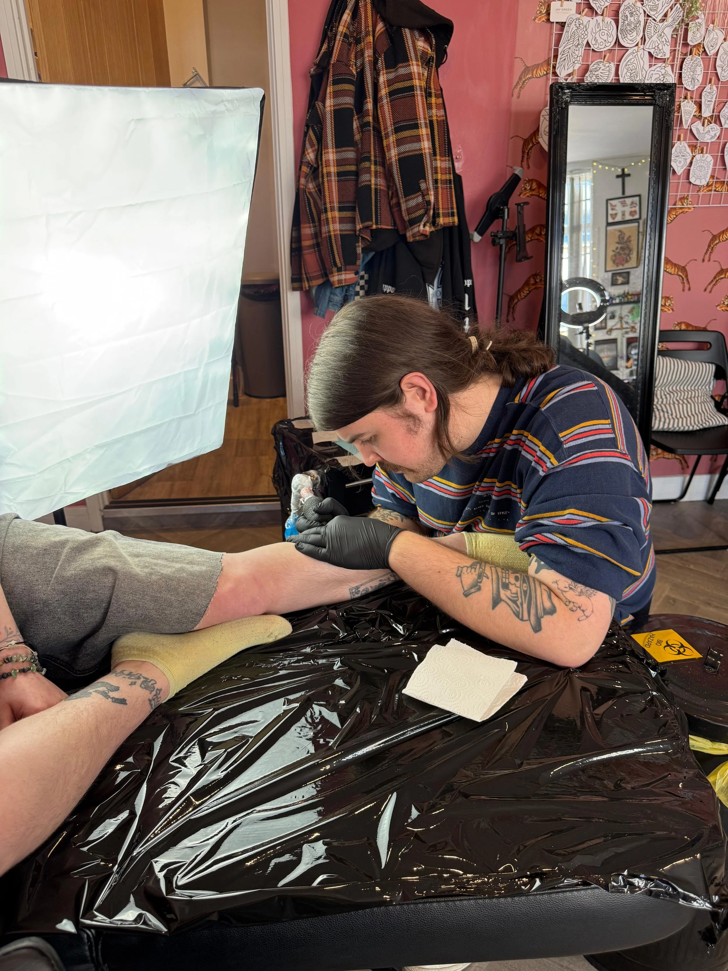 A person with long hair, tattoos, and wearing a striped shirt is tattooing a client's arm in a tattoo shop. The client is lying on a table covered with a black plastic sheet, with their arm extended. The shop has pink walls, a large mirror, and various decorations including a wall grid with paper cutouts and a cross. Tattoo equipment and supplies are visible on the table.