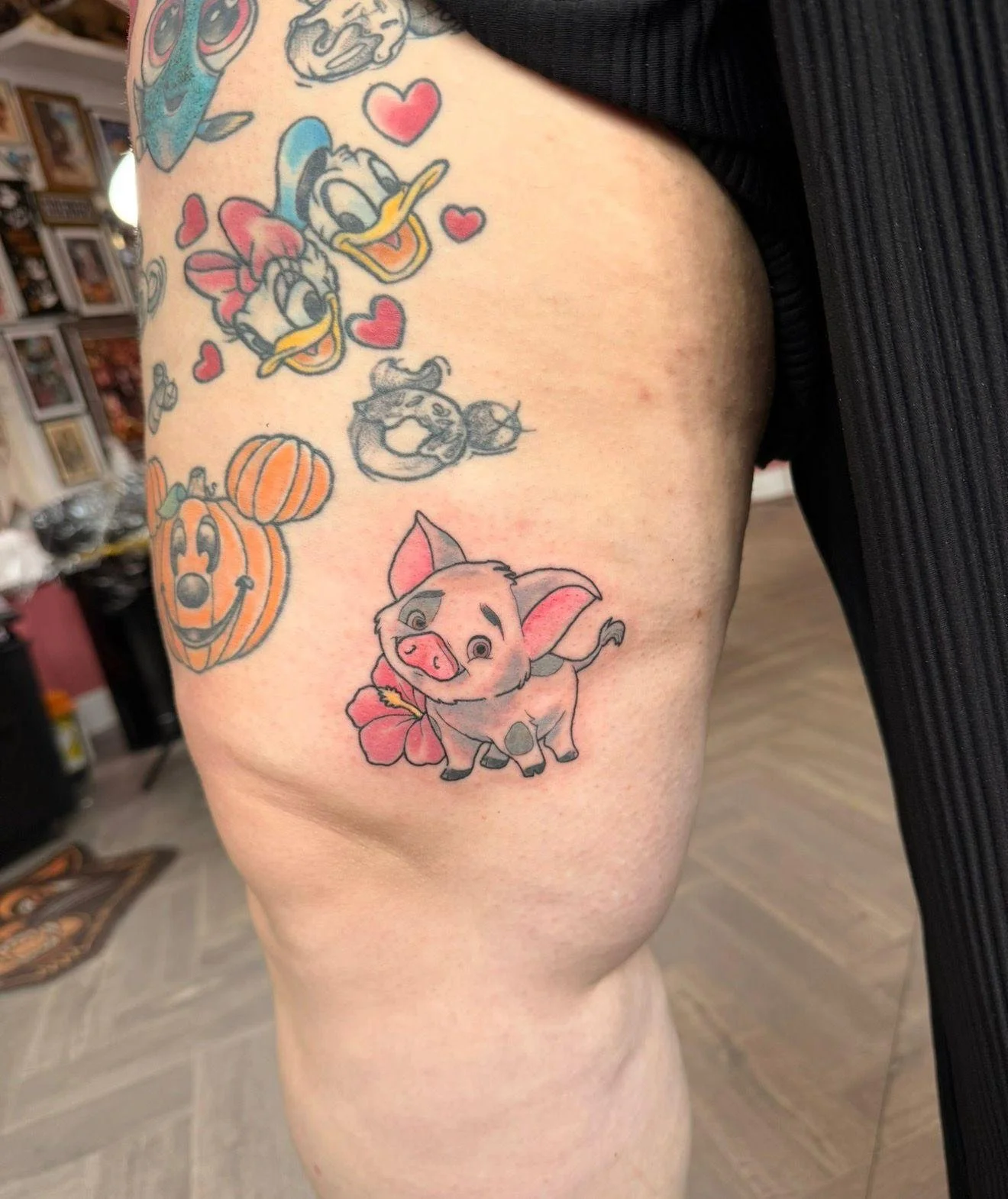 Tattoo of Disney characters Donald Duck, Daisy Duck, and a piglet with hearts, pumpkins, and flowers on a person's upper thigh.