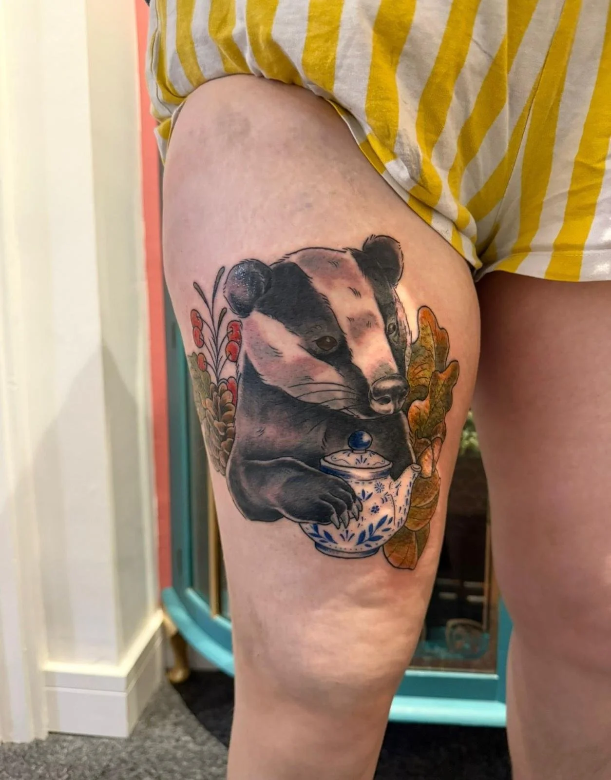 Tattoo of a badger holding a porcelain sugar bowl, surrounded by leaves and red berries, on a person's thigh.