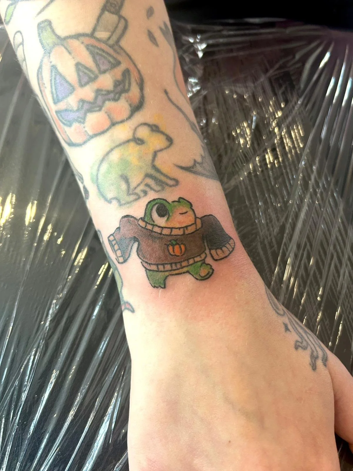 Tattoo of a cute green cartoon turtle wearing a large brown sweater with a small pumpkin on it, on a person's forearm.