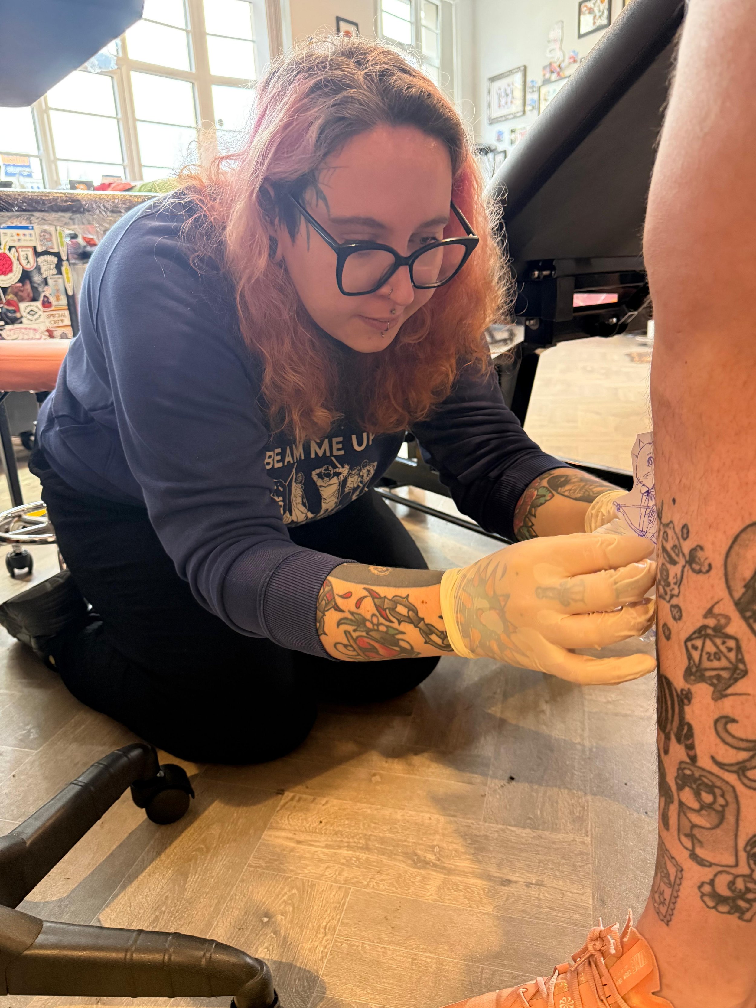 A woman with red hair, black glasses, and tattoos on her arms, kneeling on the floor in a tattoo studio, is tattooing a person's leg.
