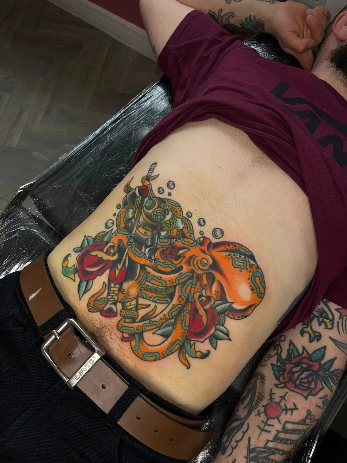 A colorful tattoo of a stylized orange octopus with intricate blue and black patterns, surrounded by red roses with green leaves, on a person's side ribcage.