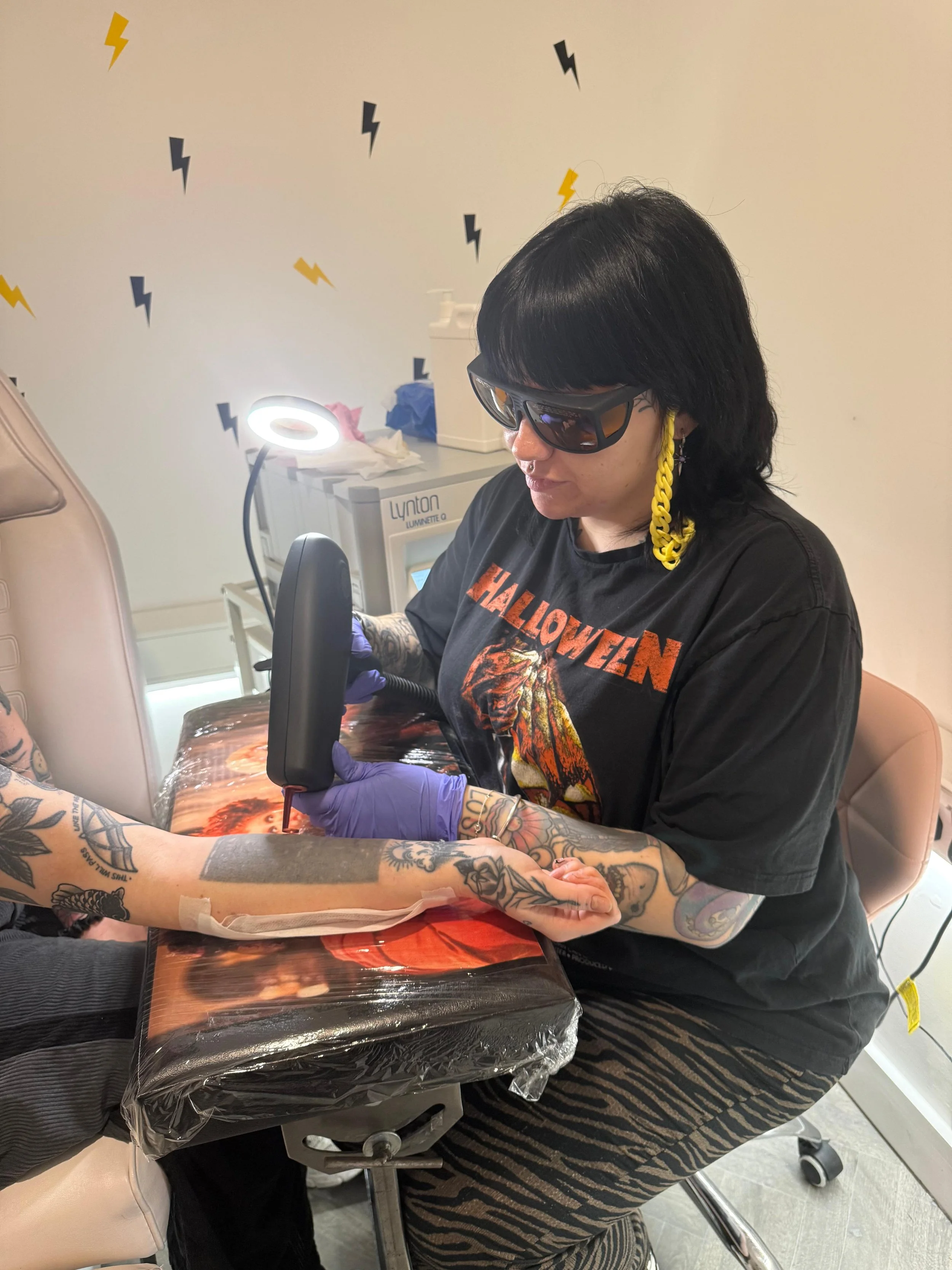 A person with black hair with bangs wearing sunglasses, a black T-shirt with orange graphics, and a yellow chain earring, tattooed arms, and purple gloves, is getting a tattoo on their forearm from a tattoo artist in a tattoo studio.