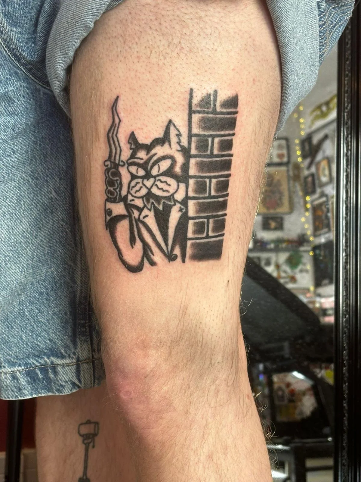 A tattoo on a person's upper thigh showing an anthropomorphized cat holding a lightsaber, with half of a brick wall behind it.