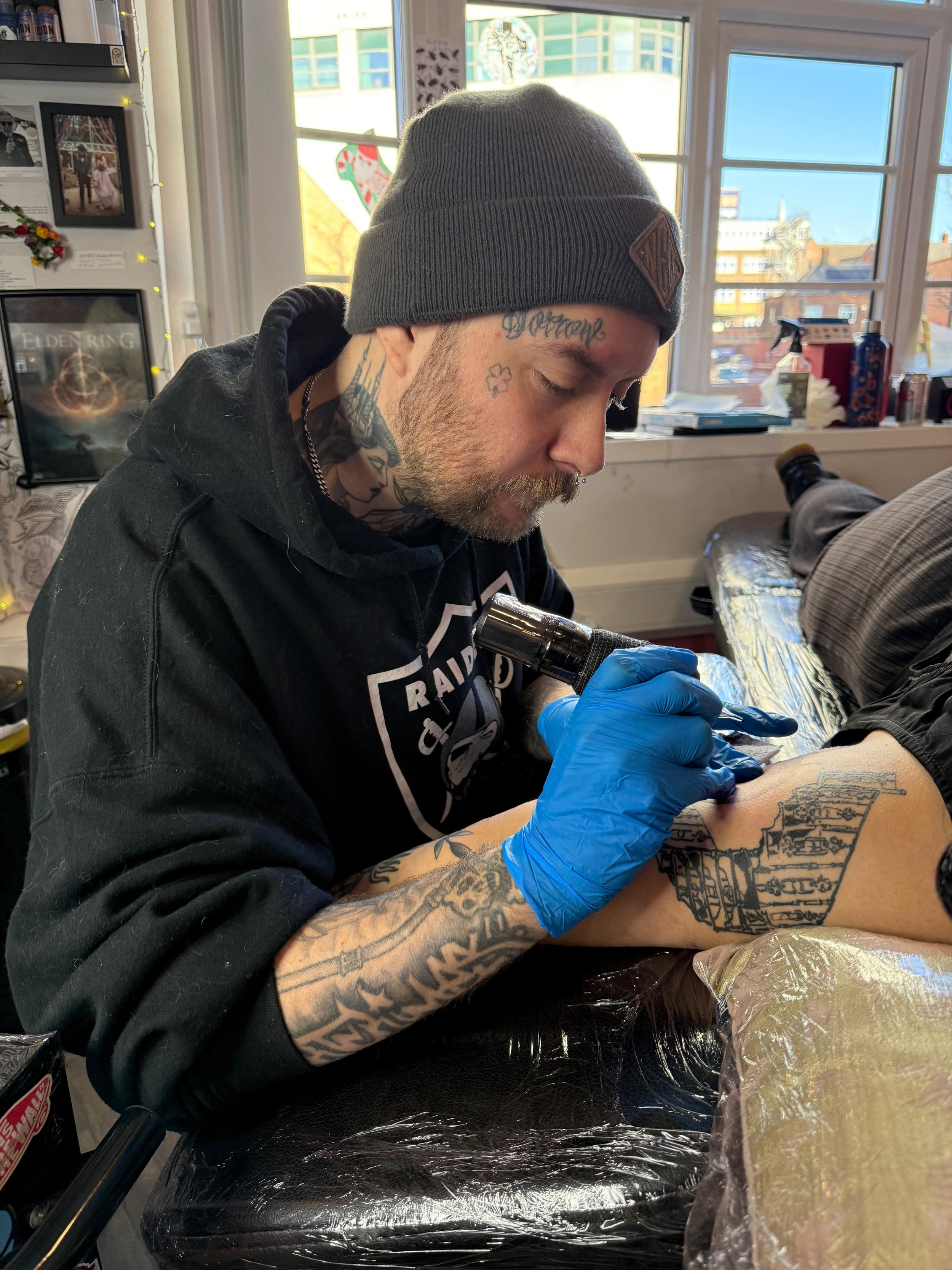 A man with tattoos on his face and arms getting a tattoo on his arm in a tattoo studio. He is wearing a gray beanie and a black hoodie, and has a moustache and beard. The tattoo artist is wearing blue gloves and is working on the man's forearm.