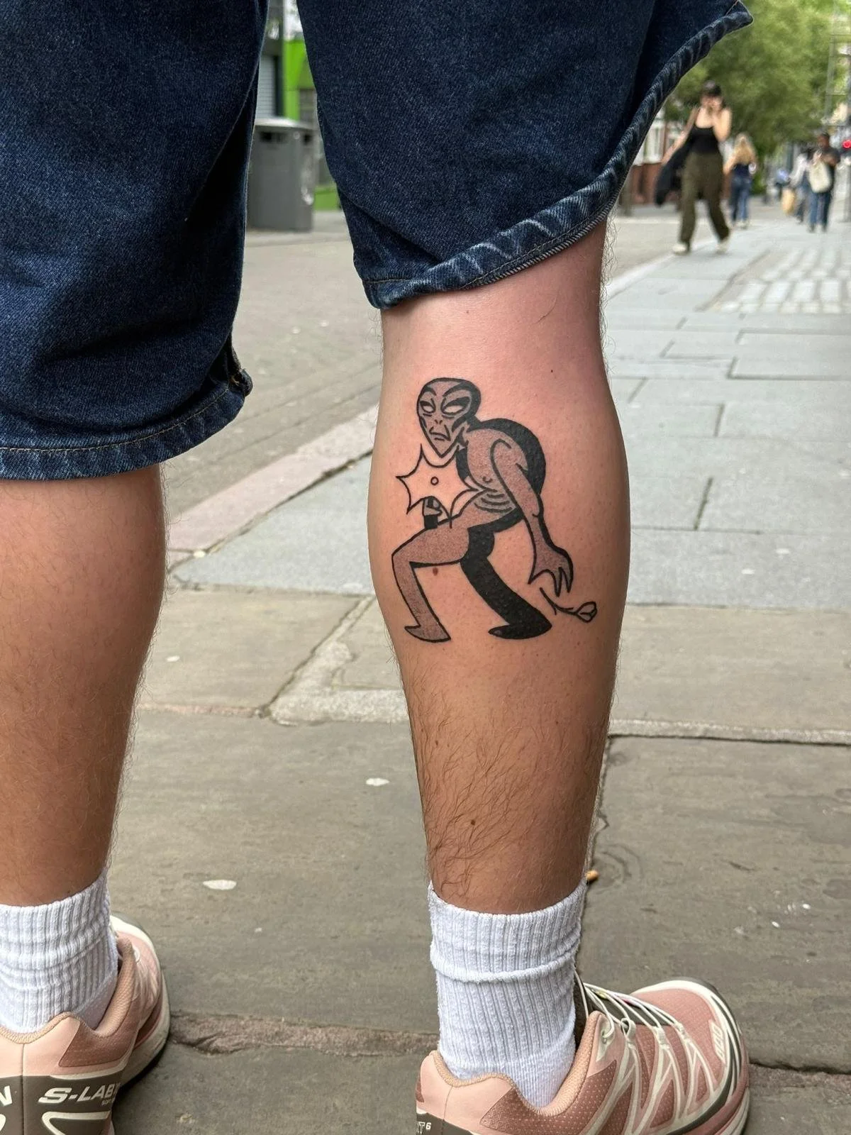Tattoo of a cartoonish alien with large eyes and a stern expression, crouching with bent knees, on a person's right calf. The person is wearing denim shorts, white socks, and pink and beige sneakers, standing on a city sidewalk.