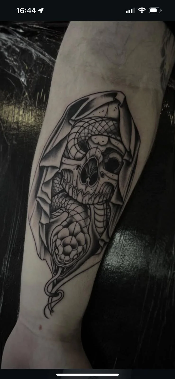 Black and gray tattoo of a skull inside a hood, combined with snake elements and a chain pattern on the forehead.