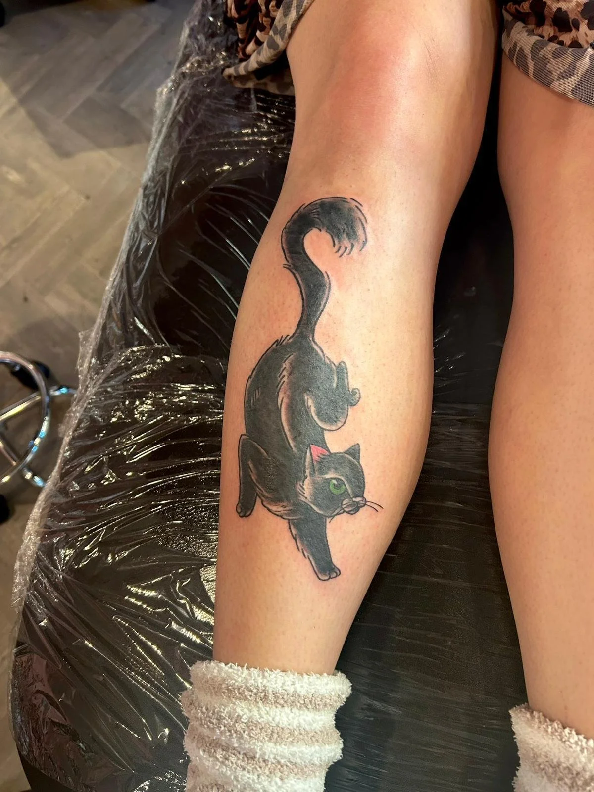 A tattoo of a black cat with green eyes, shown in a stretching pose with its tail curved upward, on a person's leg.