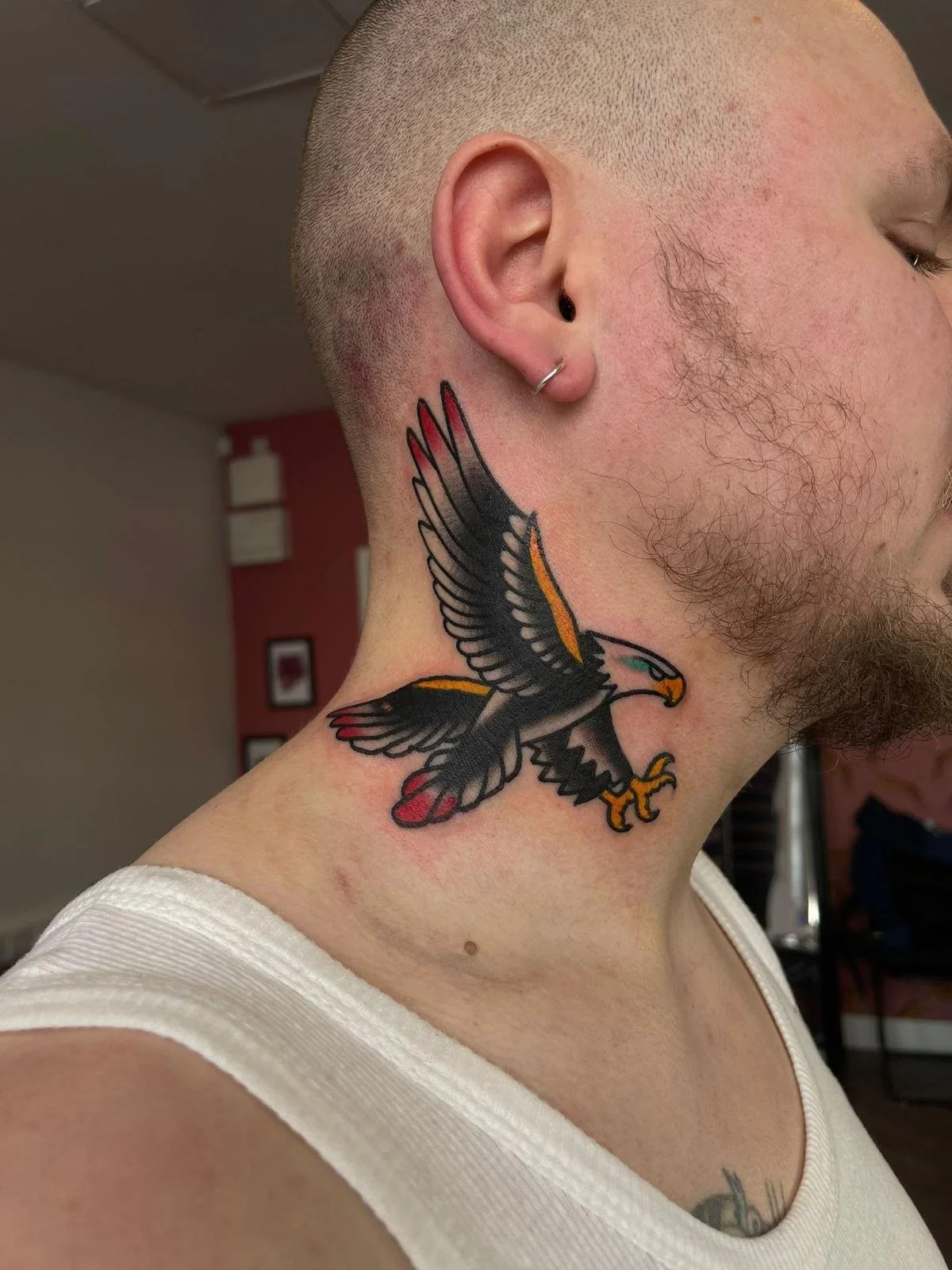 A close-up of a person's neck and face showing a colorful eagle tattoo, with the person having a shaved head, a small hoop earring, and a beard.