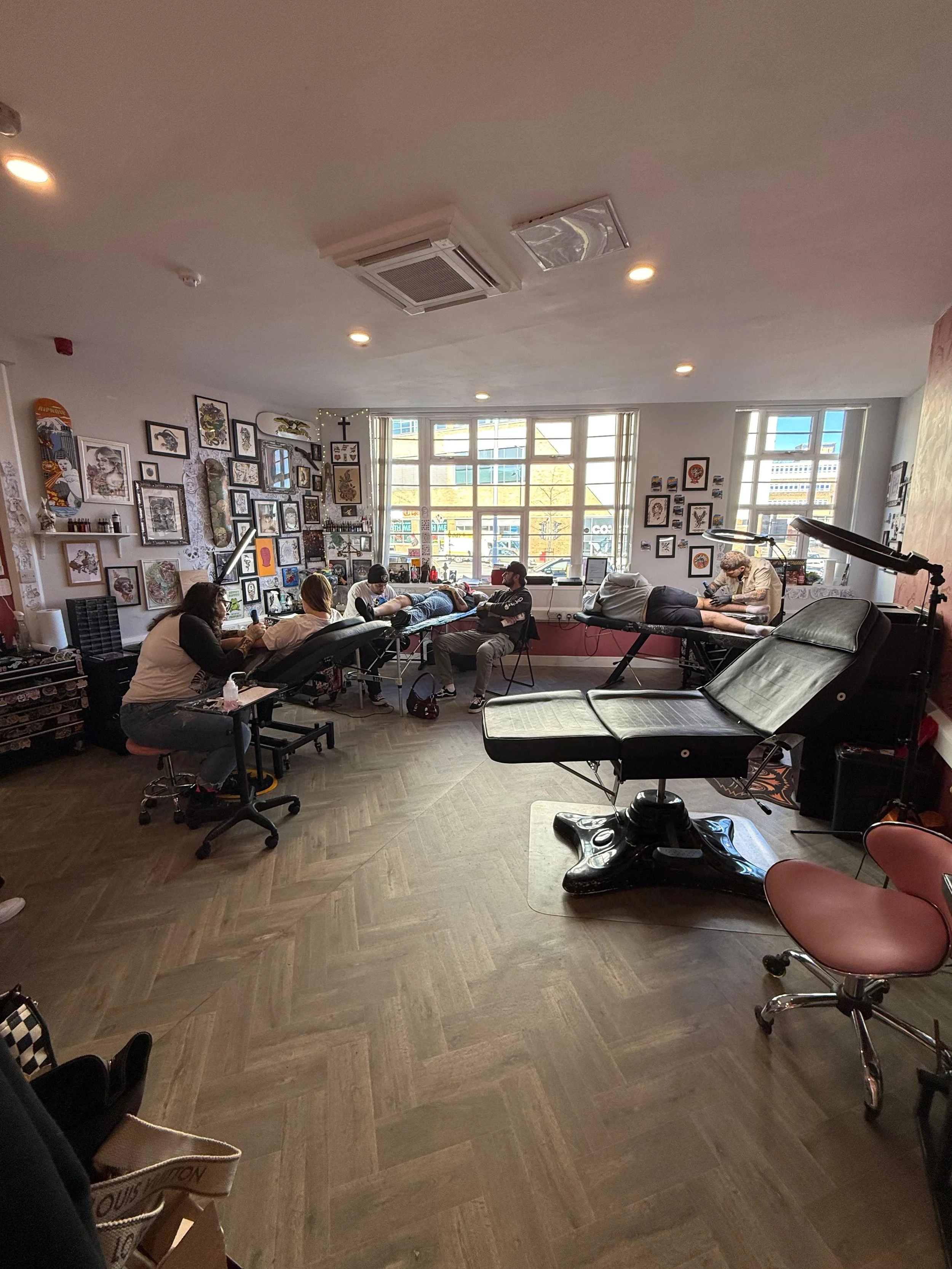 Tattoo studio with clients, tattoo artists, and artwork on the walls, in a well-lit room with large windows.