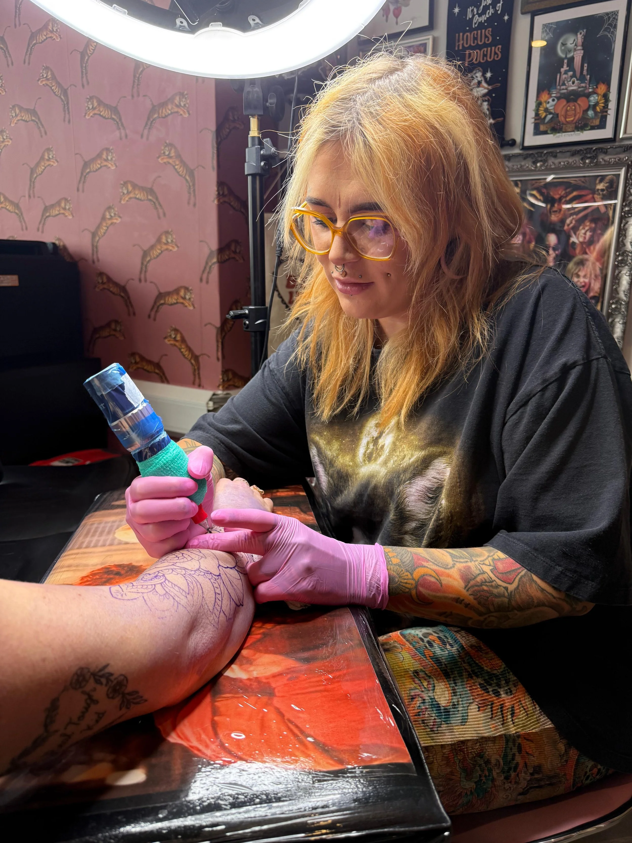 A tattoo artist with glasses and colorful tattoos is tattooing someone's arm in a tattoo studio with Halloween-themed posters and tiger wallpaper in the background.