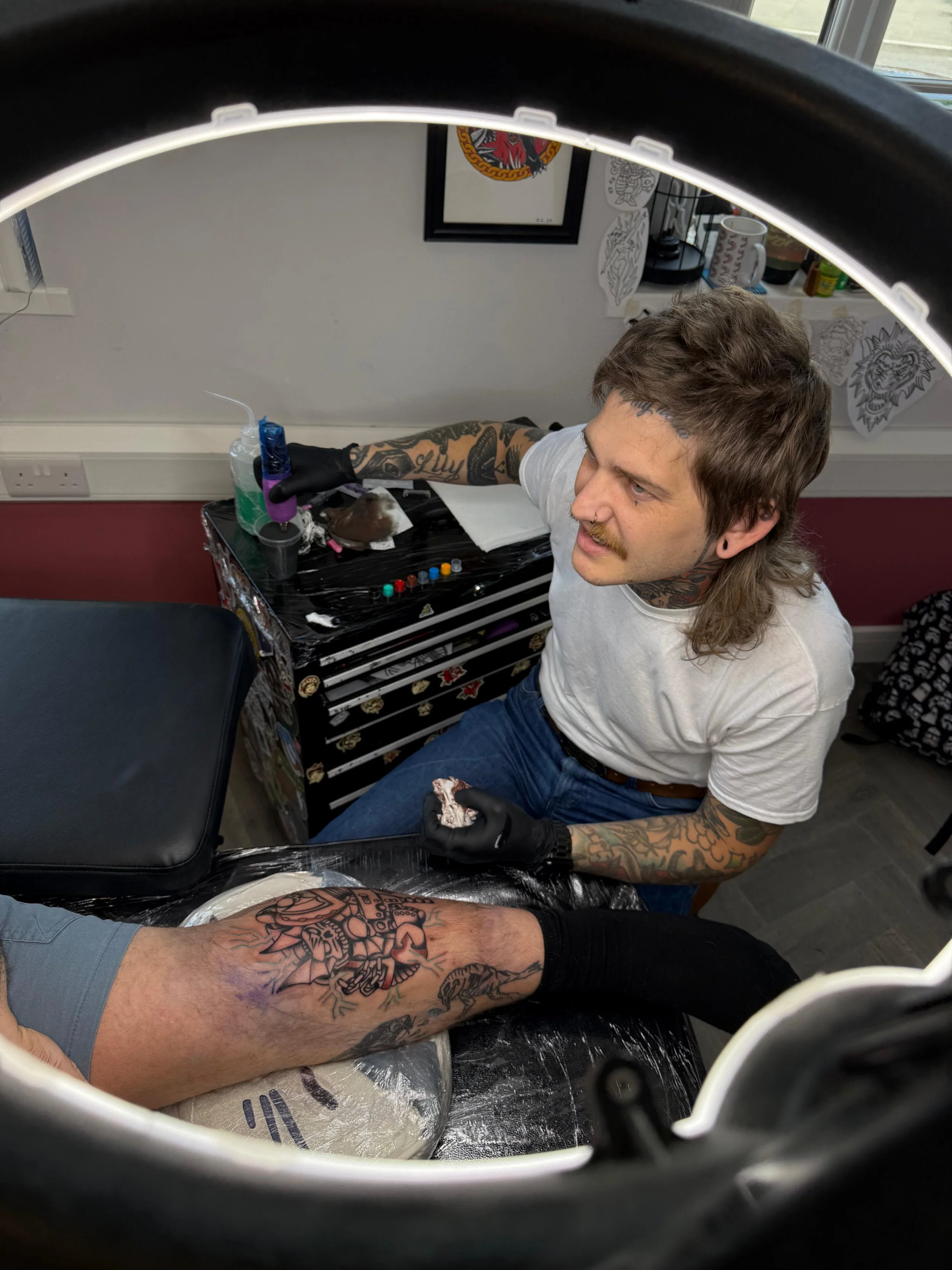 A man getting a tattoo on his thigh inside a tattoo studio. The photo is taken from inside a tattoo helmet, showing the artist with tattoos on his arms, wearing gloves, working on the man's leg.