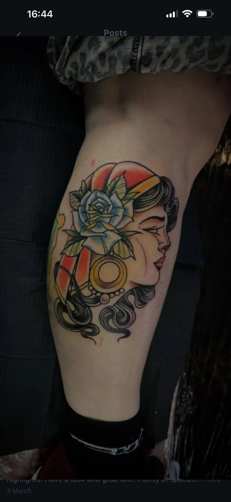 Tattoo of a woman’s face in profile with flowing hair, a blue rose, and jewelry with a sunset background.