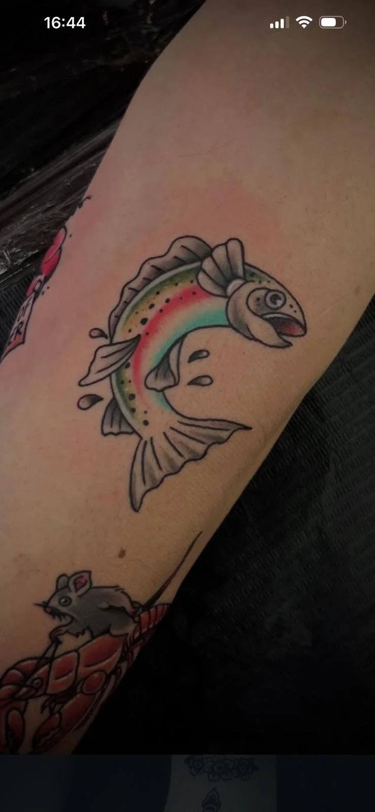 Tattoo of a cartoon-style fish with rainbow colors on its body, jumping out of water droplets.