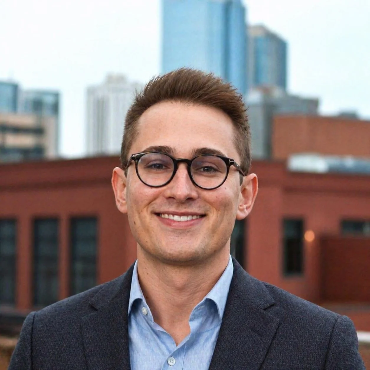 A smiling young man with glasses and short brown hair, wearing a blue shirt and dark blazer, standing outdoors with city buildings in the background.