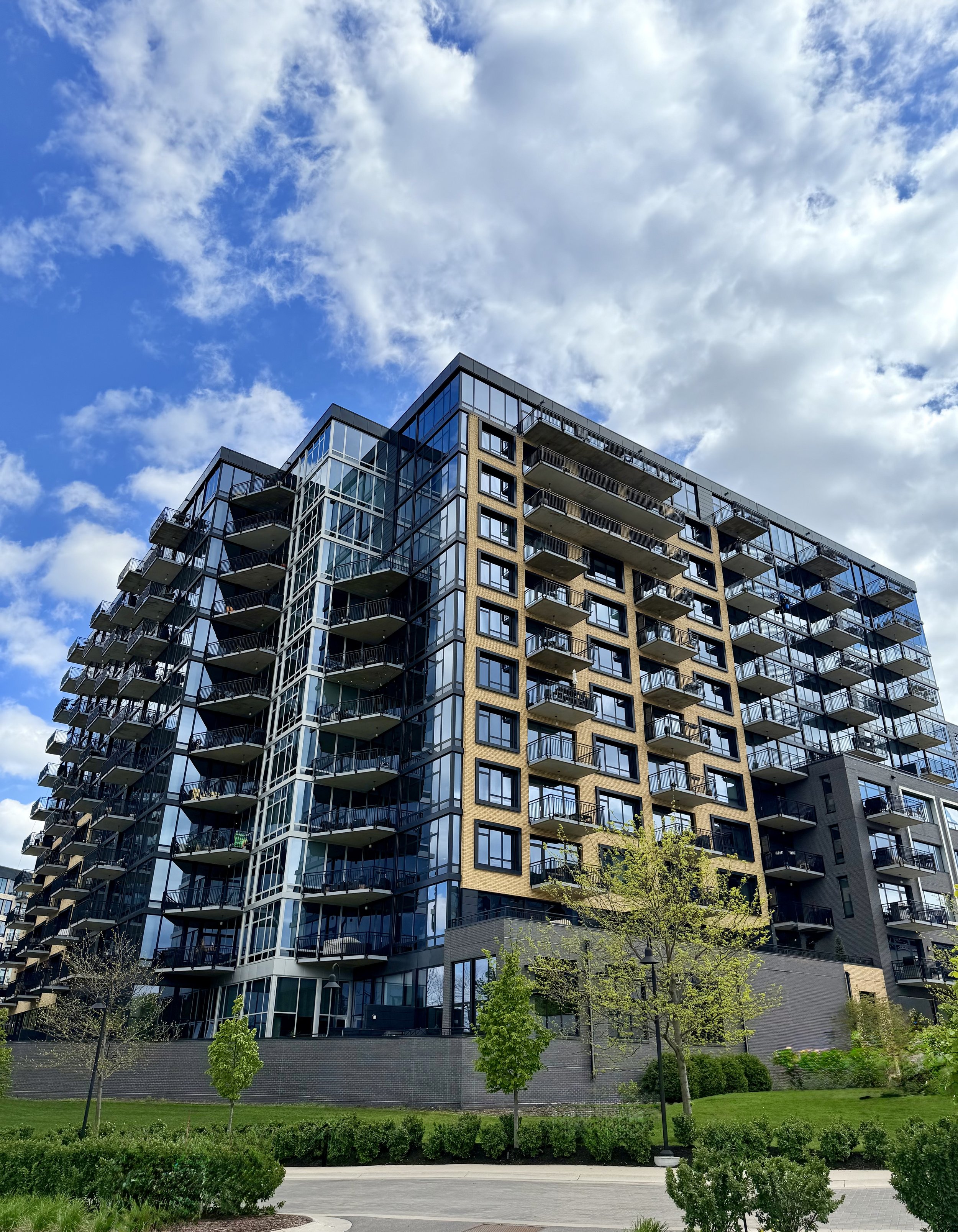 A modern multi-story residential building with glass balconies, set against a partly cloudy sky, with a landscaped foreground featuring green trees and bushes.
