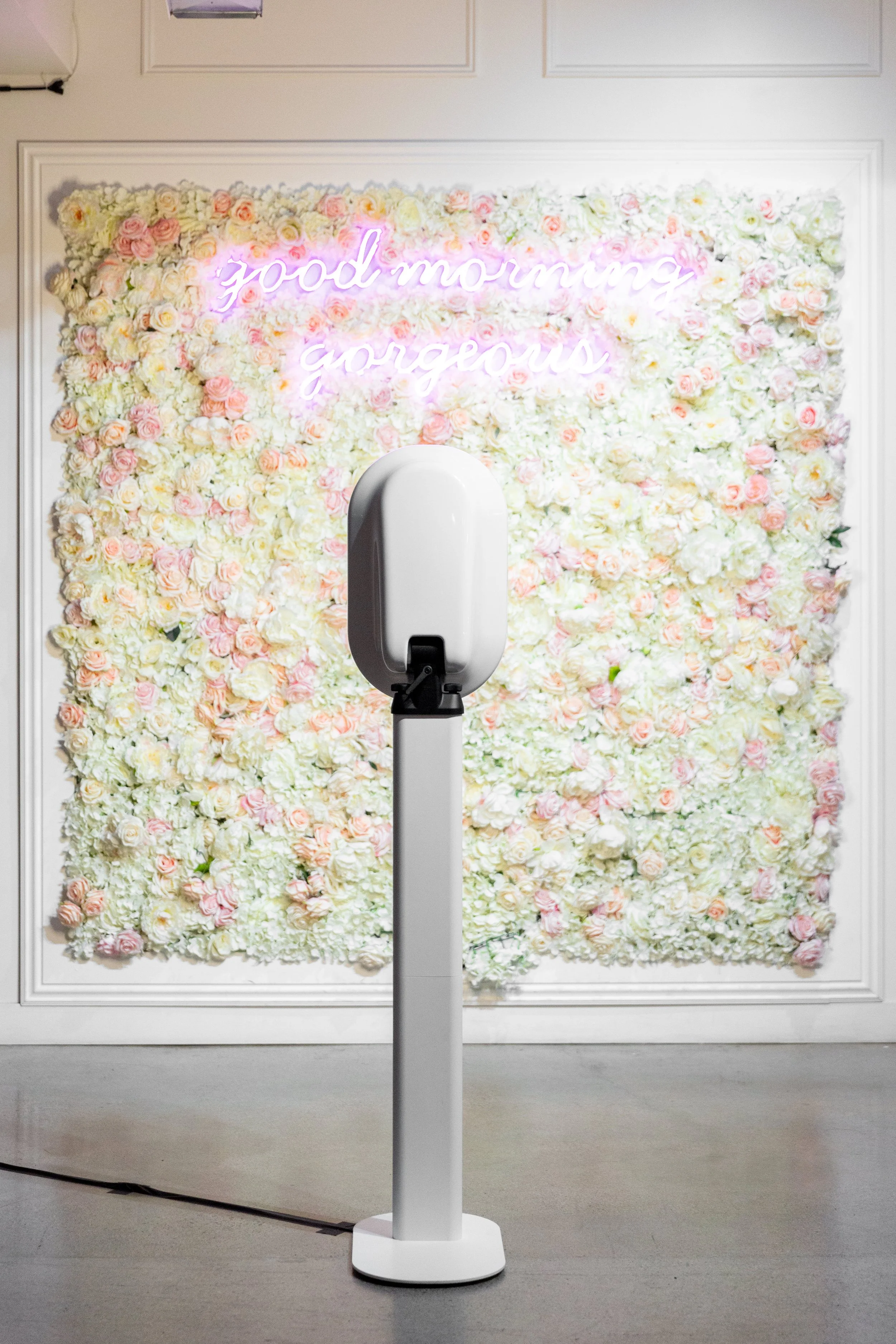 A floral wall with pink and white roses and a neon sign that says 'good morning gorgeous,' with an object resembling a hand sanitizer dispenser in front of it.